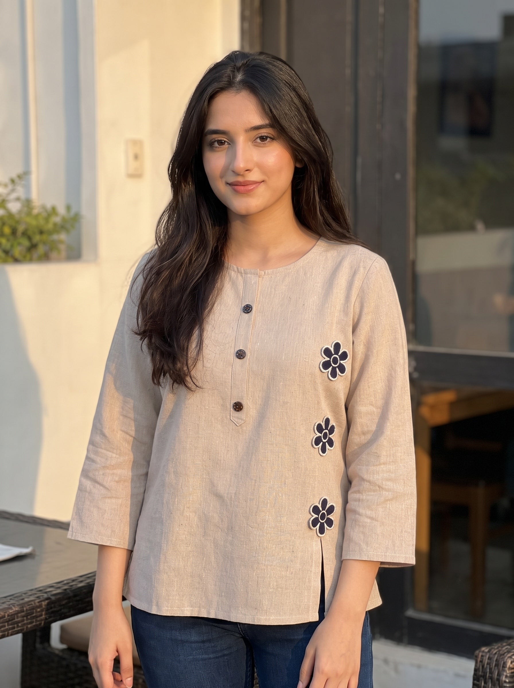 A woman wearing a light beige linen three-quarter sleeve top with dark buttons and floral embroidery, paired with dark blue jeans, standing in front of a modern building background.