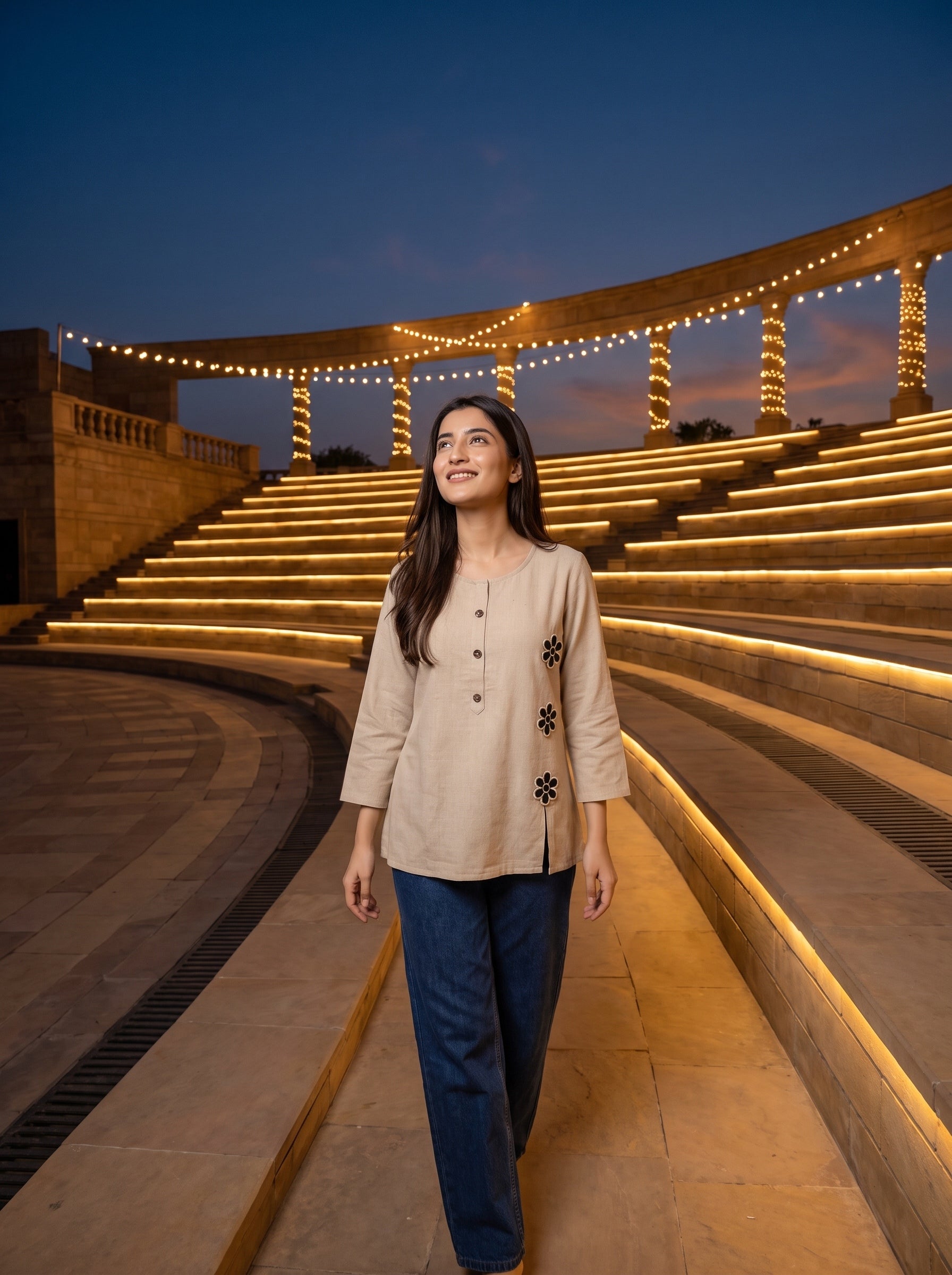 A woman wearing a beige long-sleeve tunic with black floral embroidery and dark blue jeans walks through a modern outdoor amphitheater at dusk, with illuminated stone steps and columns in the background.