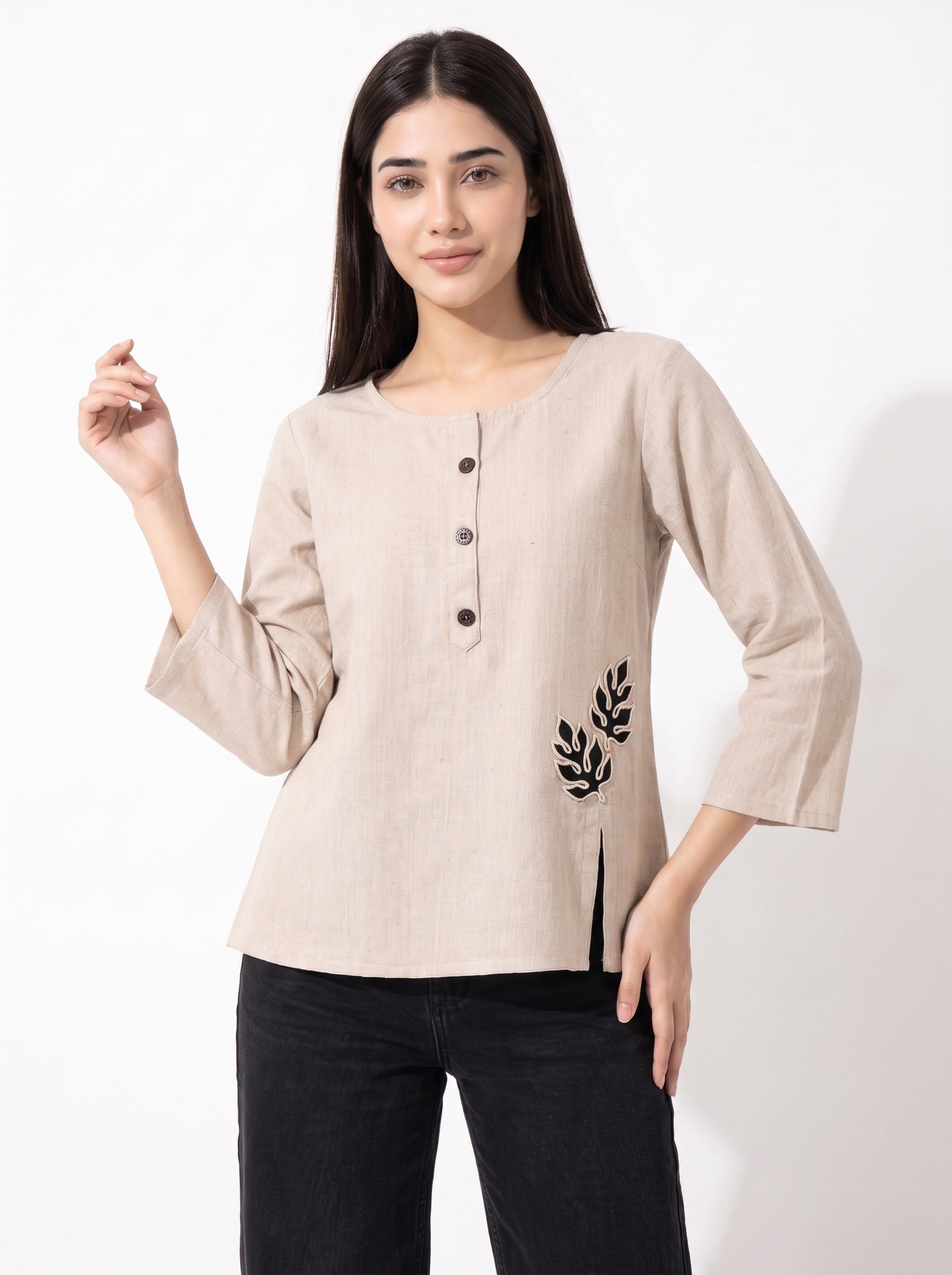 A woman modeling a light beige linen long-sleeve top with three-quarter sleeves, a buttoned neckline, and black leaf embroidery on the lower left side, paired with black pants against a plain white background.