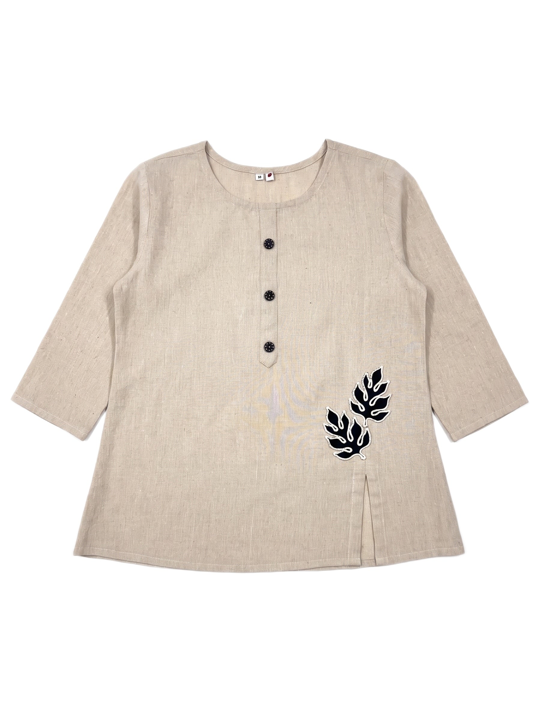 A beige linen long-sleeve tunic top with a three-button henley neckline and black embroidered leaf motifs on the lower right side, displayed flat against a white background.