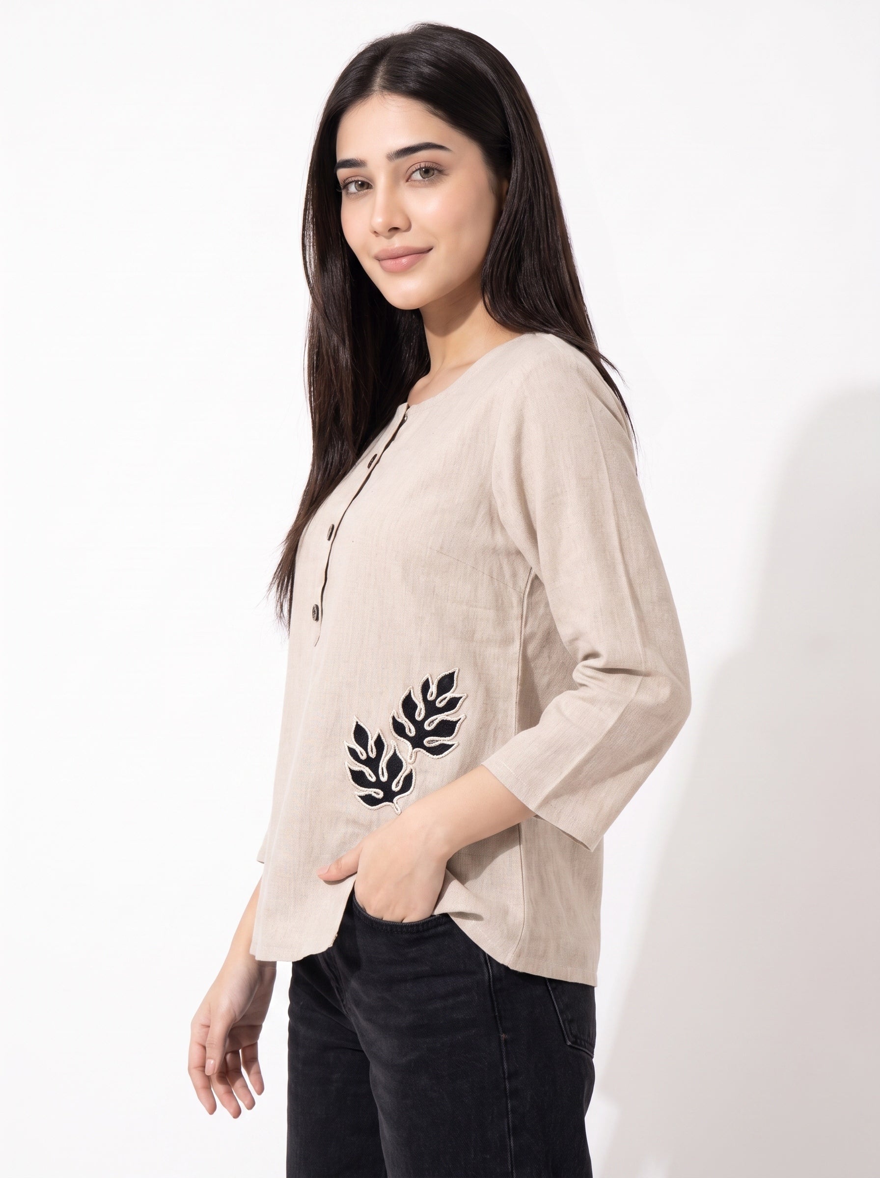 A woman modeling a light beige linen three-quarter sleeve top with black leaf embroidery on the front, paired with black pants, against a plain white background.