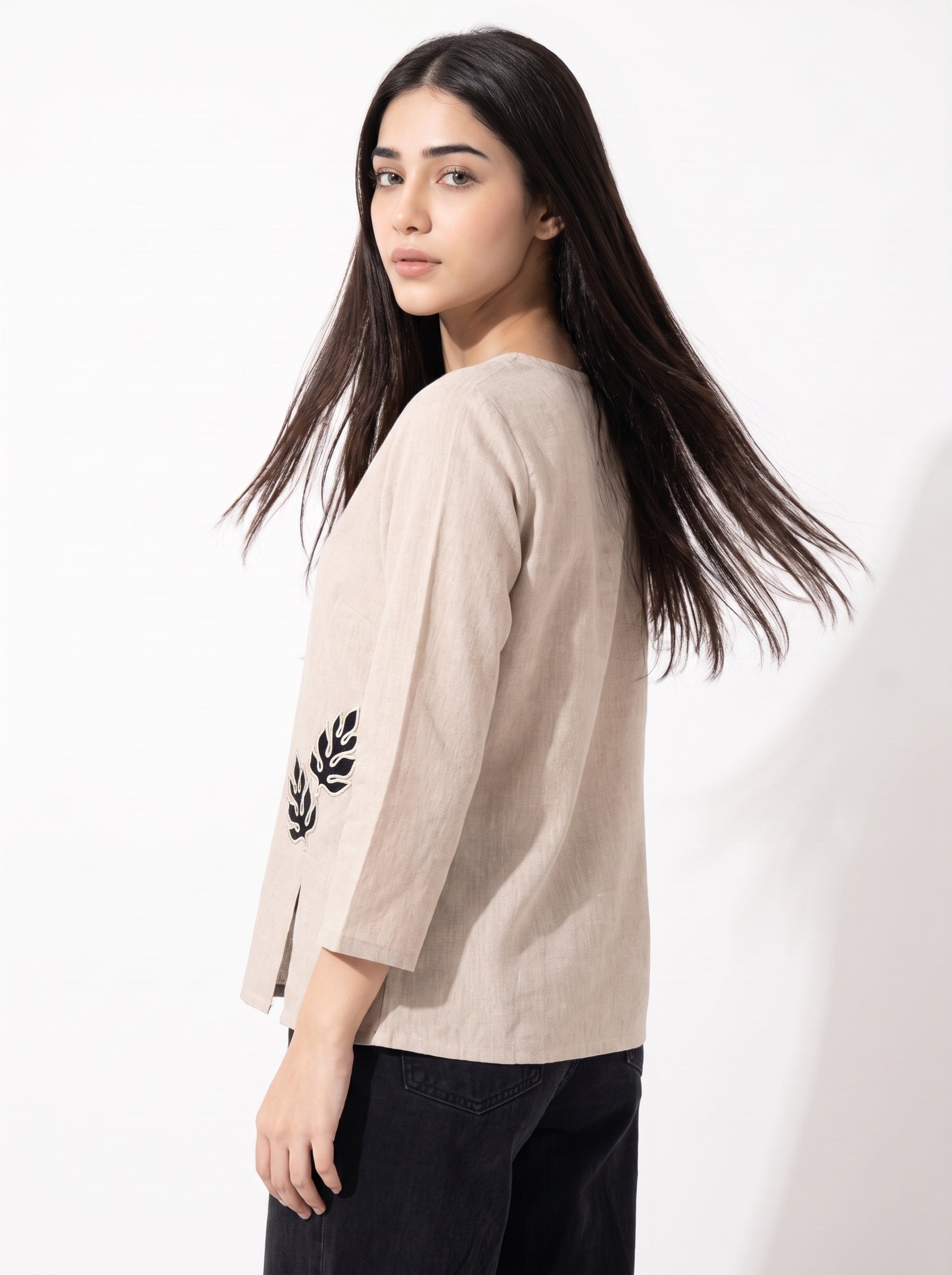 A woman with long dark hair is shown from a side-back angle wearing a light beige linen three-quarter sleeve top with black embroidered leaf detailing on the sleeve, paired with black pants, against a plain white background.