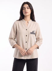 A woman modeling a light beige linen button-up shirt with a stand collar, black buttons, and a small embroidered graphic on the left chest. The shirt has three-quarter sleeves and is paired with black pants against a plain white studio background.
