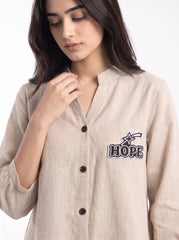 A woman wearing a light beige linen button-up shirt with a dark embroidered 'HOPE' logo on the left chest, set against a plain white background.
