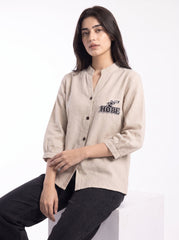 A woman wearing a beige linen button-up shirt with a black embroidered 'HOPE' graphic on the chest, paired with black jeans, seated against a white studio background.