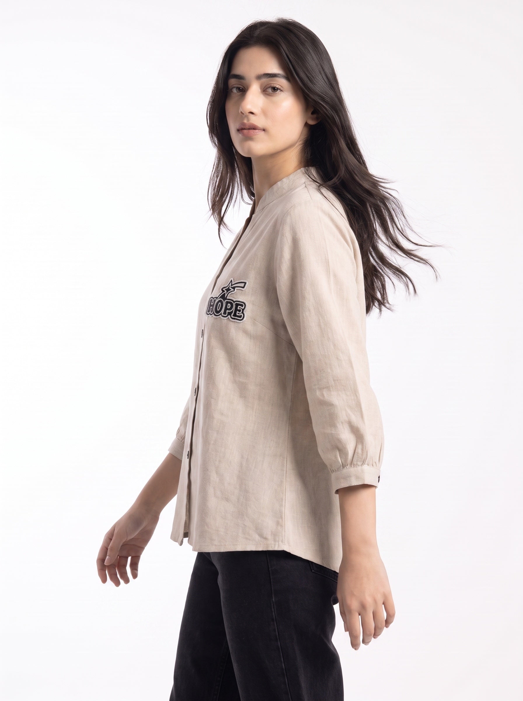 A woman modeling a light beige linen button-up shirt with black embroidered lettering on the chest, paired with black pants, against a plain white background.