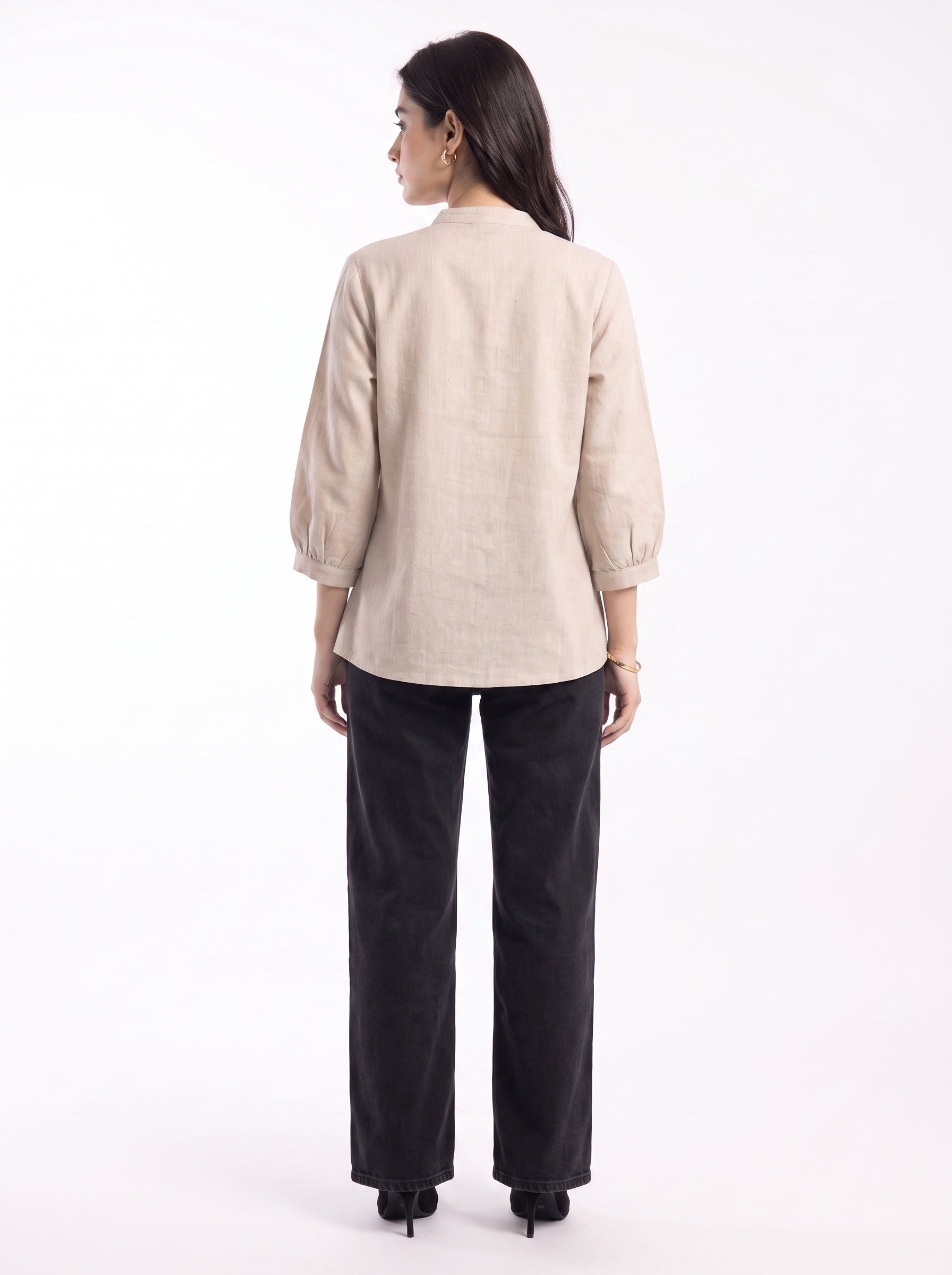 A woman is shown from the back wearing a light beige linen three-quarter sleeve blouse and black wide-leg trousers, standing against a plain white studio background.