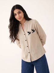 A woman modeling a beige linen button-up shirt with black embroidered birds on the chest, paired with blue jeans, against a plain off-white background.