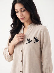 A woman wearing a light beige linen button-up shirt with two black embroidered birds on the chest, photographed against a clean white background.
