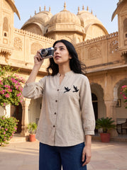 A woman stands in a courtyard in front of a domed sandstone building, holding a vintage camera to her eye. She is wearing a light beige linen button-up shirt with black embroidered birds on the chest and dark blue jeans.