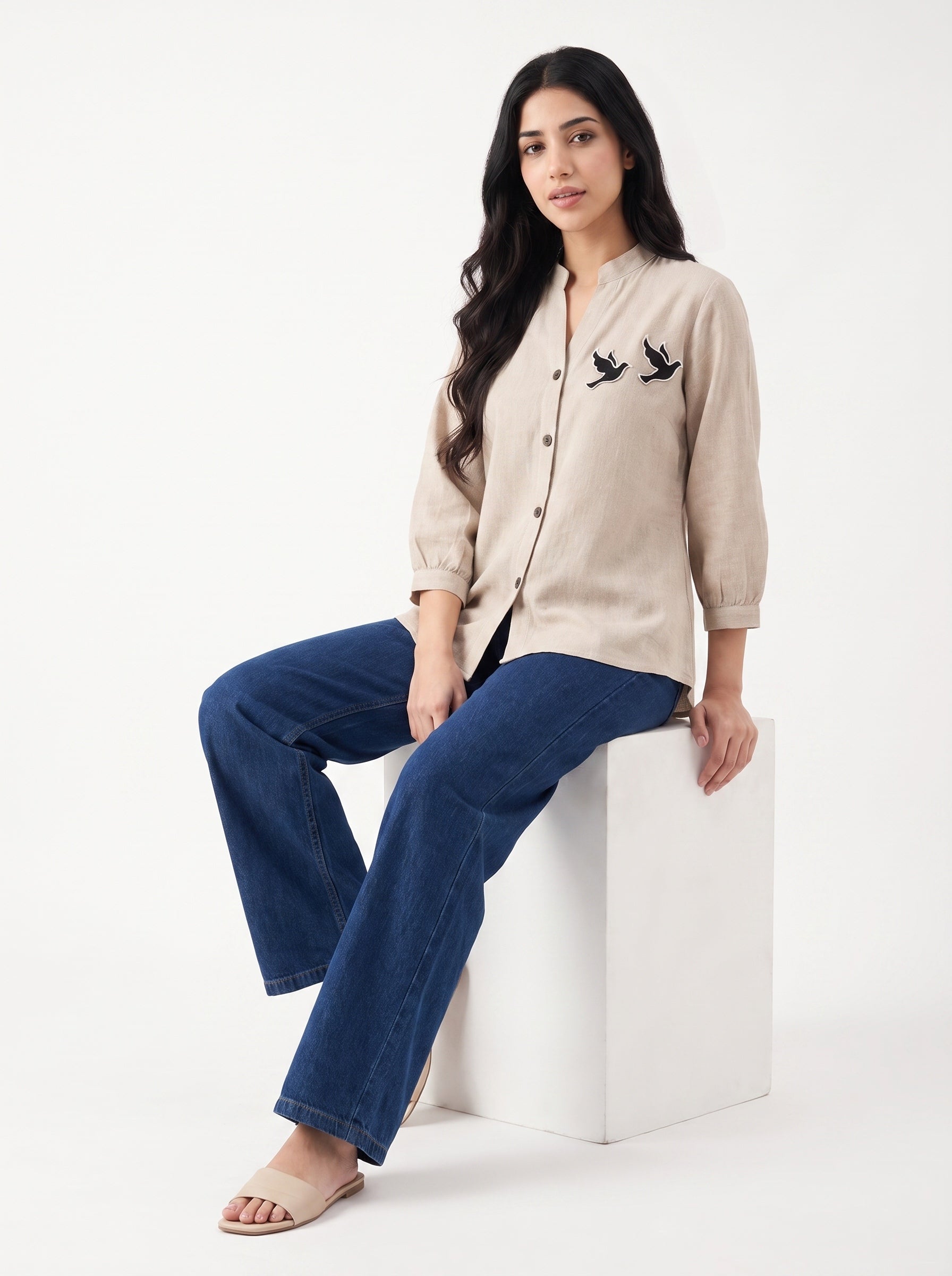 A woman sits on a white cube in a studio setting, wearing a beige linen button-up shirt with black bird embroidery on the chest and dark blue jeans.
