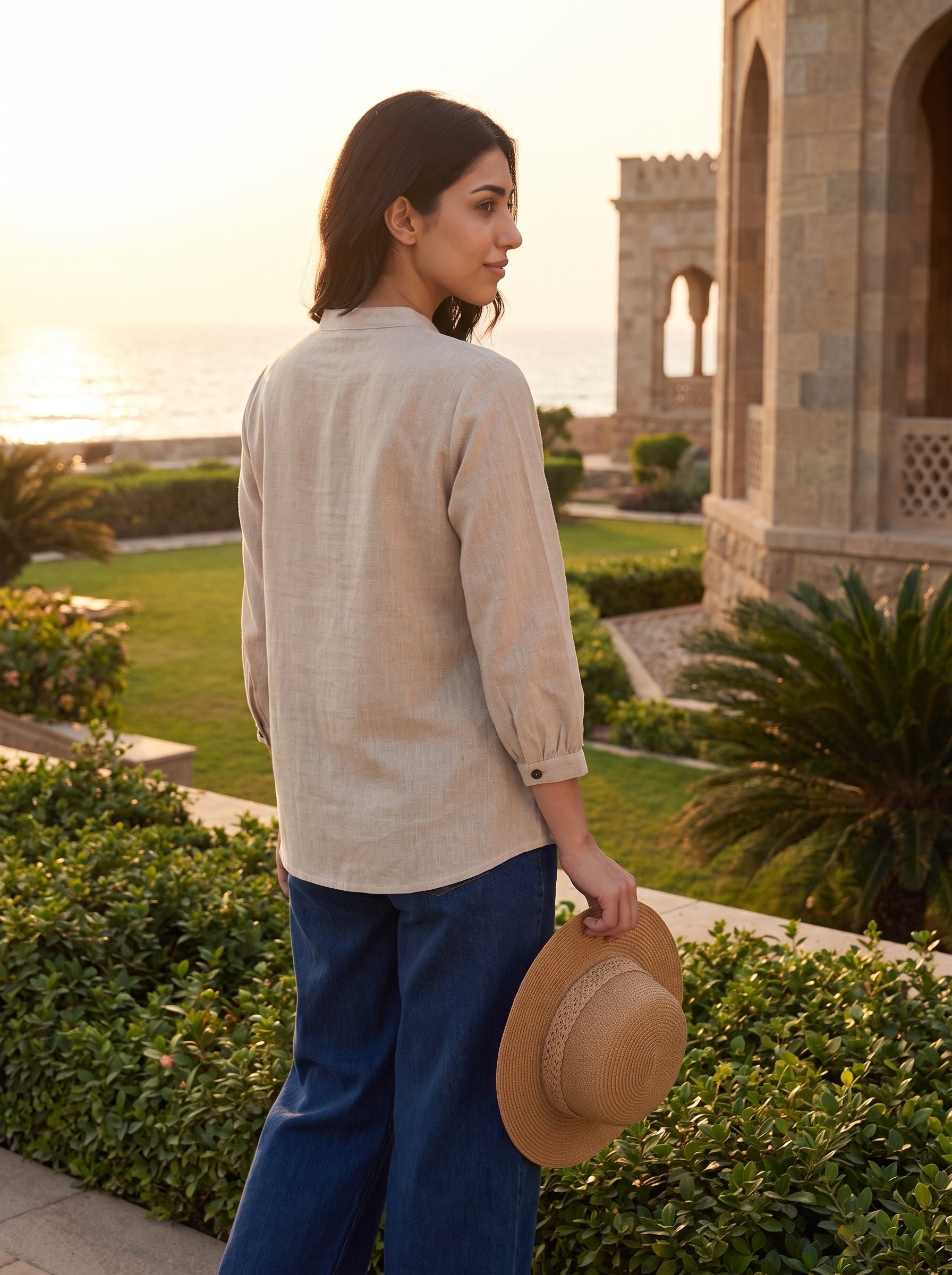 A woman stands outdoors in a garden at sunset, shown from the back wearing a light beige linen shirt and blue pants while holding a straw hat.