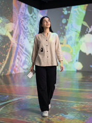 A woman walks on a glossy floor with colorful projected light patterns in the background, wearing a light beige linen button-up shirt with a black embroidered bird motif on the pocket, paired with black wide-leg pants and white sneakers.