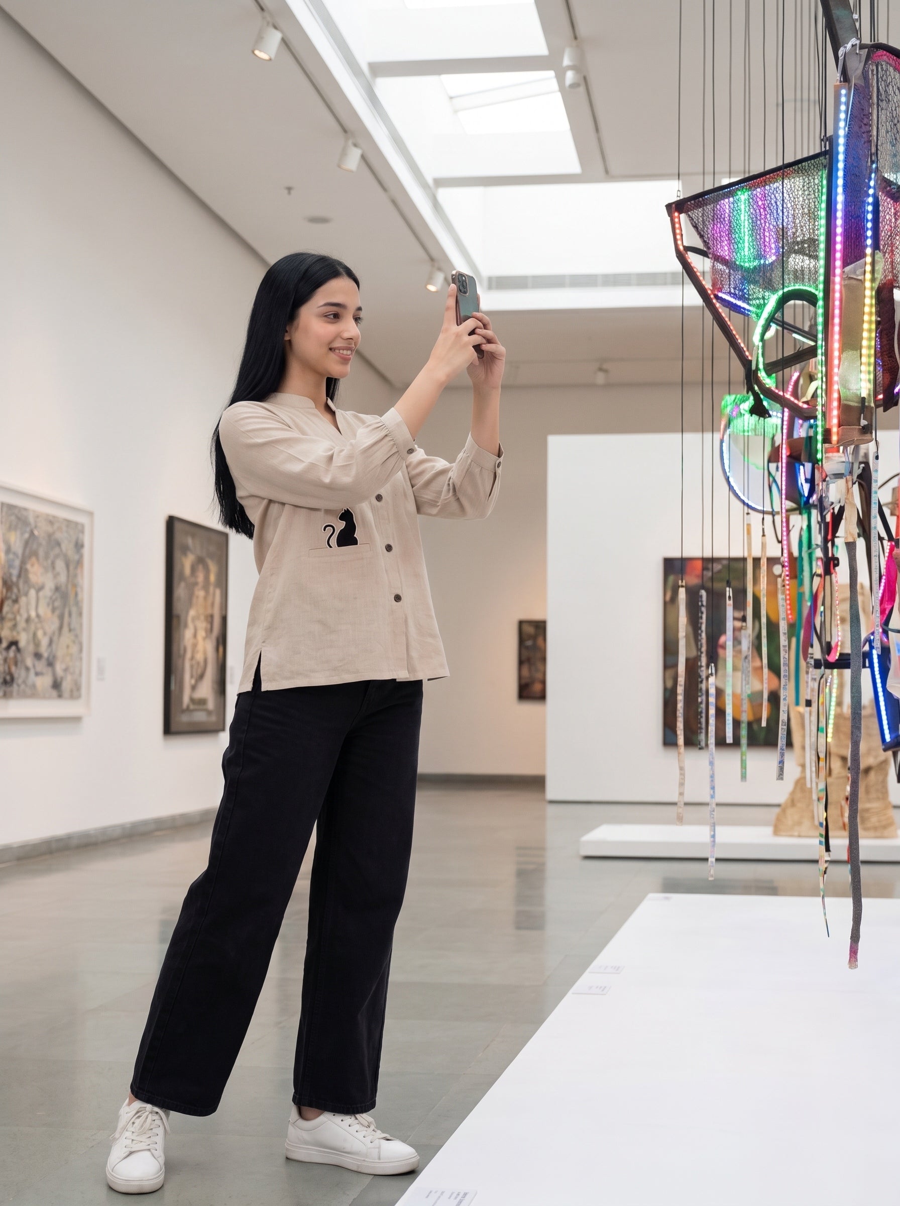 A woman in a beige button-up shirt and black wide-leg pants takes a photo of a colorful hanging textile art piece in a modern gallery space with white walls and framed paintings.