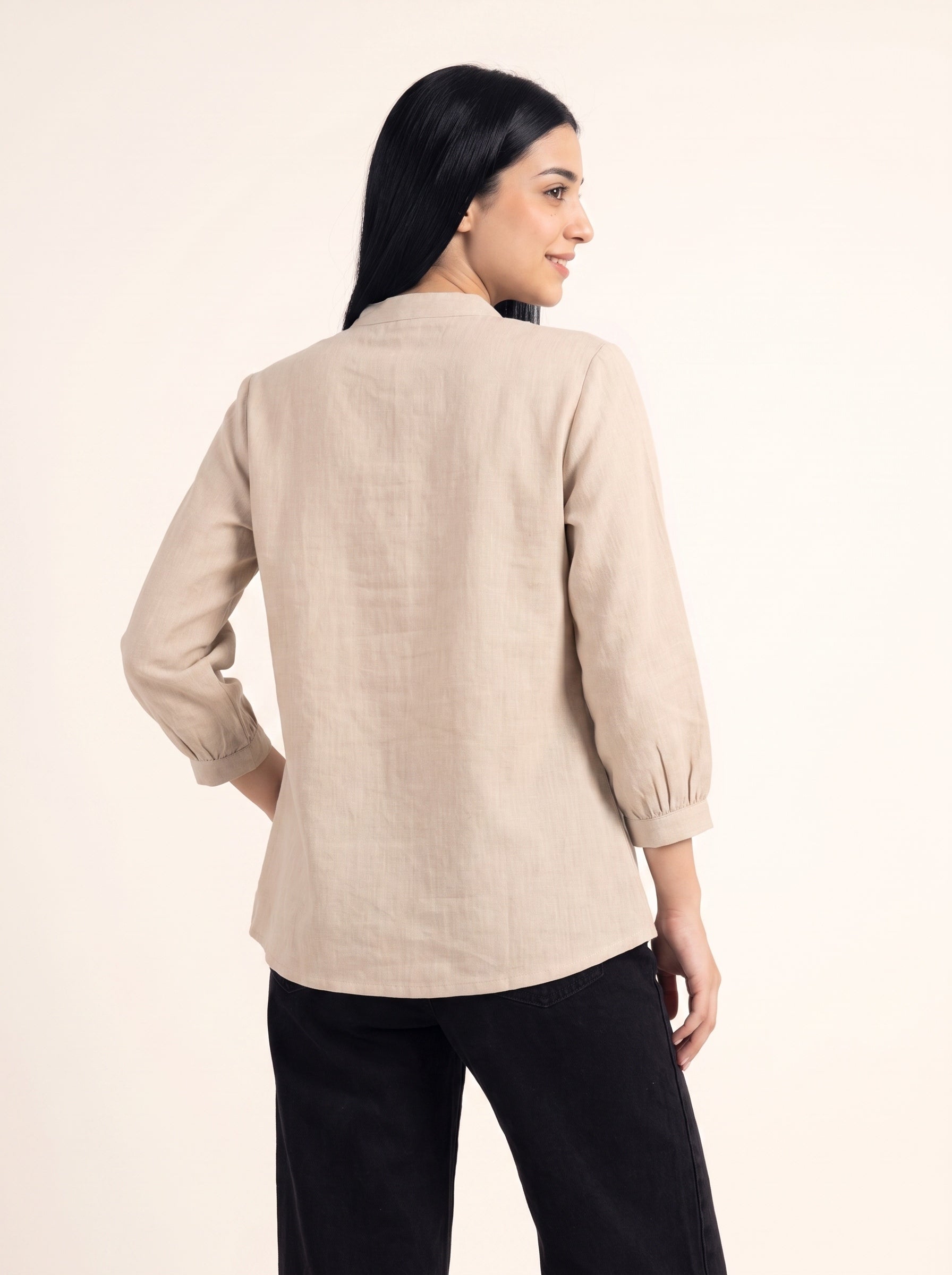A woman shown from the back wearing a light beige linen long-sleeve blouse with gathered cuffs and black pants, photographed against a plain cream studio background.