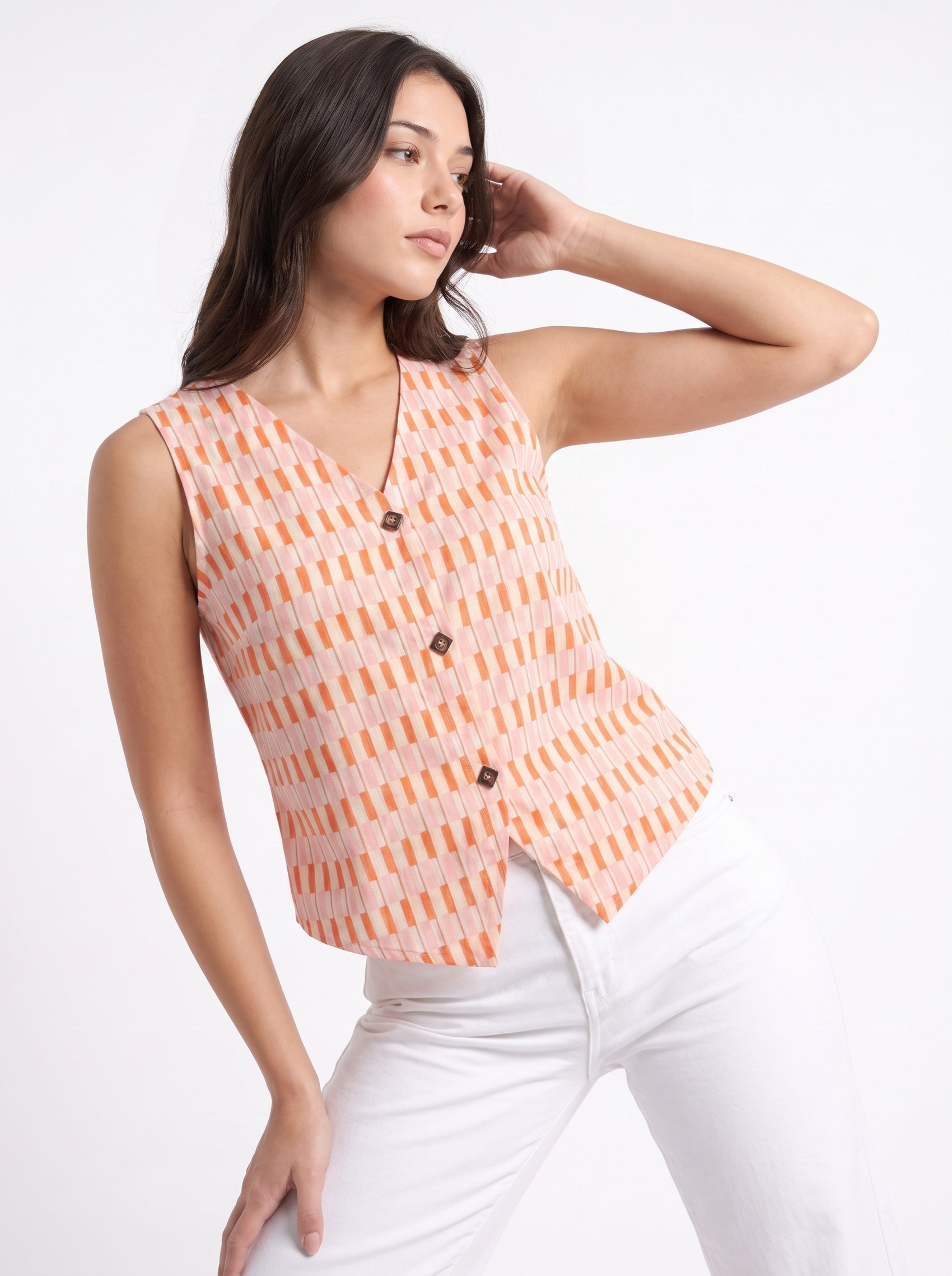 A woman modeling a sleeveless knit top with an orange and white geometric pattern, paired with white pants, against a plain white studio background.
