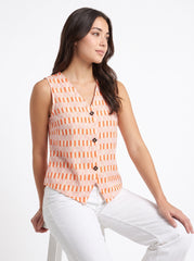 A woman wearing a sleeveless vest with an orange and white geometric pattern, paired with white pants, seated against a plain white background.