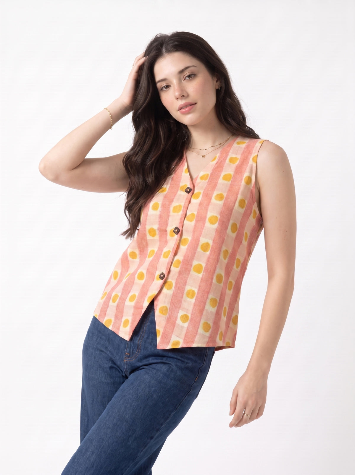 A woman modeling a sleeveless button-front vest with vertical peach and cream stripes and yellow polka dots, paired with dark blue jeans, against a plain white studio background.