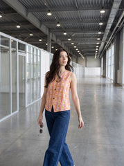A woman walking down a long, modern industrial loft hallway wearing a sleeveless coral-pink vest with a yellow and orange geometric pattern, paired with dark blue jeans.