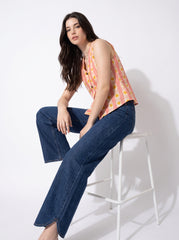 A woman sits on a white stool wearing a peach sleeveless top with yellow and orange polka dots and dark blue wide-leg jeans against a white studio background.