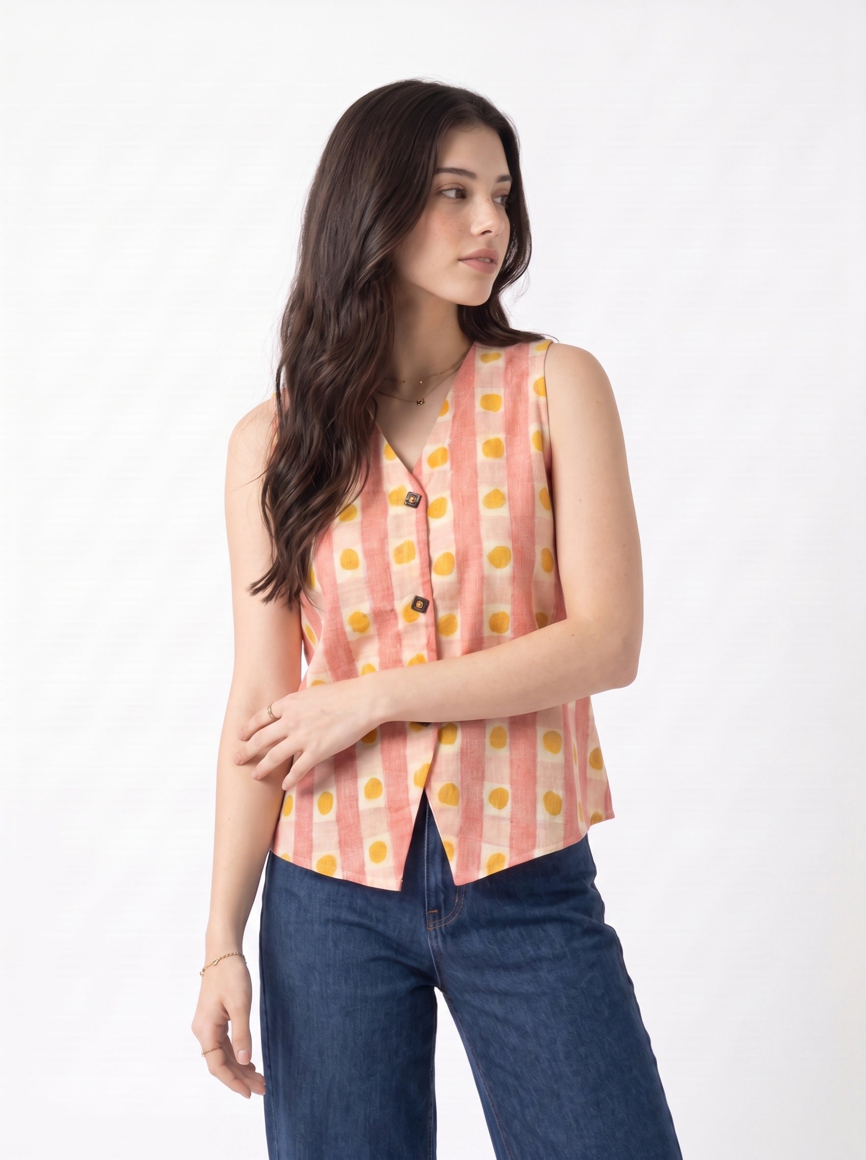 A woman modeling a sleeveless pink and orange striped vest with yellow polka dots and dark blue jeans, photographed against a plain white studio background.
