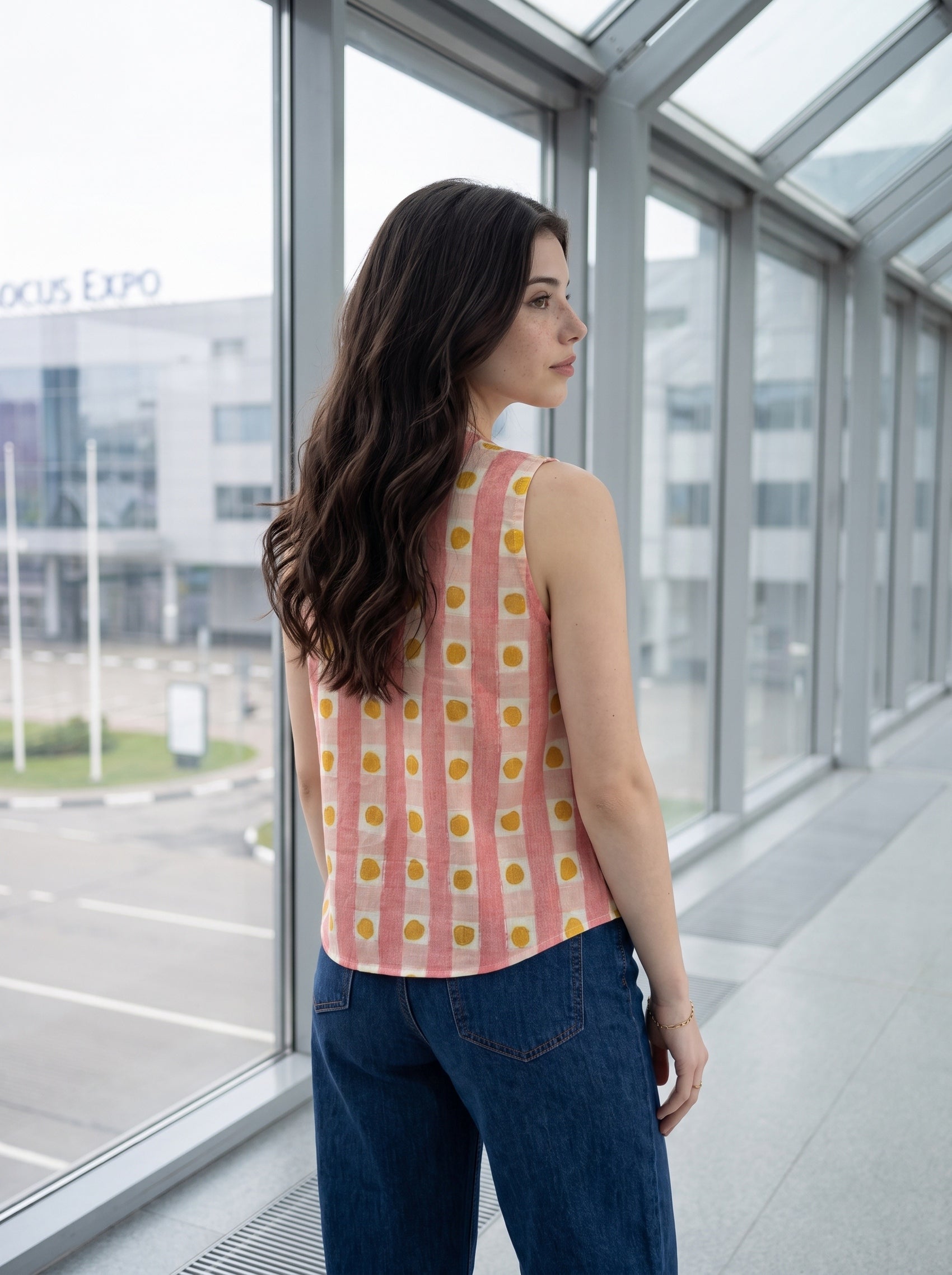 A woman with long dark hair stands in a modern glass-walled corridor, wearing a sleeveless pink top with yellow polka dots and blue jeans, looking out toward a building exterior.