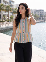 A woman stands outdoors by a waterfront wearing a sleeveless peplum-style top with vertical pastel stripes in blue, yellow, pink, and beige, paired with black pants.