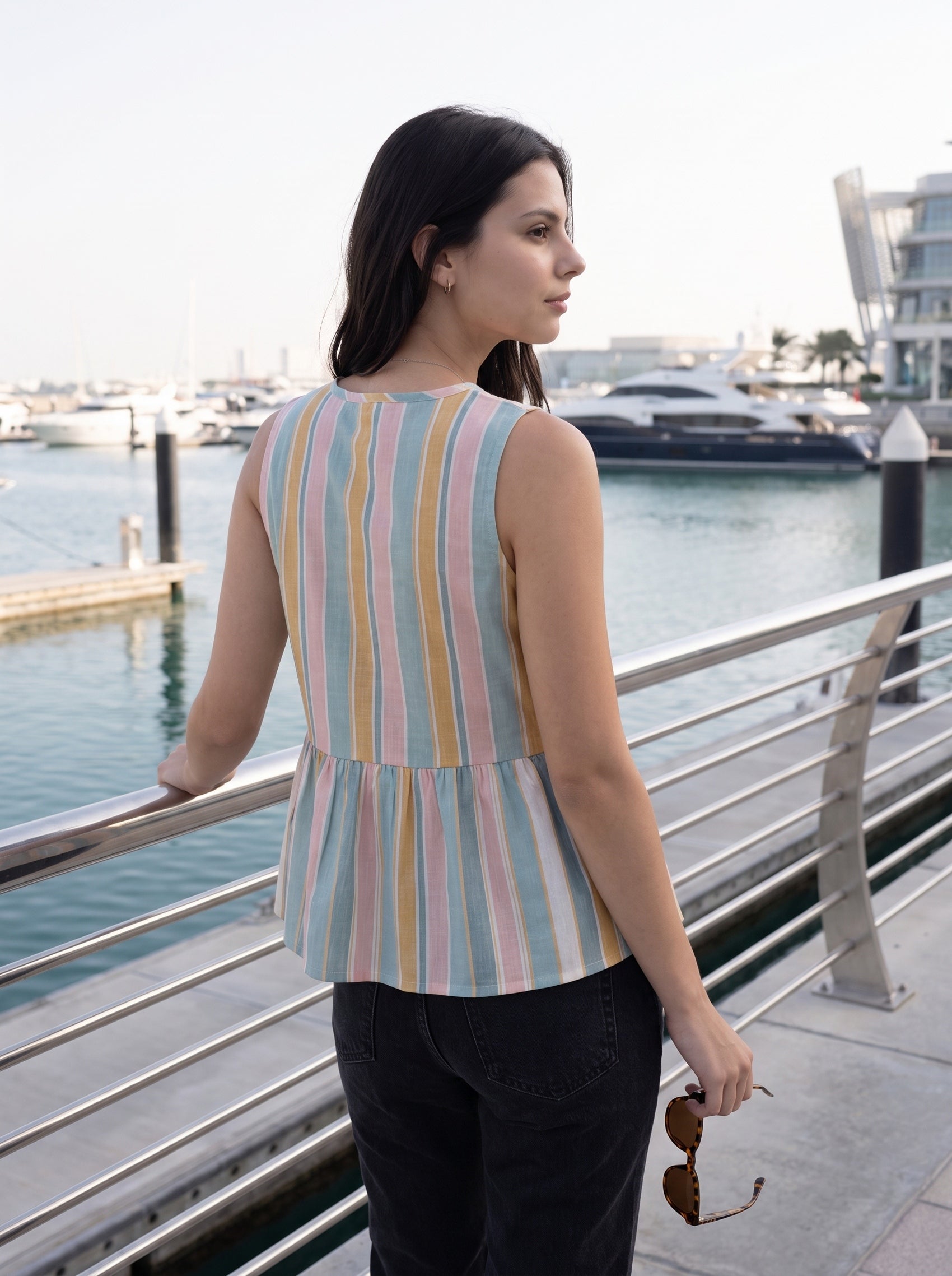A woman stands on a waterfront walkway wearing a sleeveless peplum top with pastel vertical stripes in pink, blue, yellow, and white. She is shown from the back, looking out over the water with boats and a marina in the background.