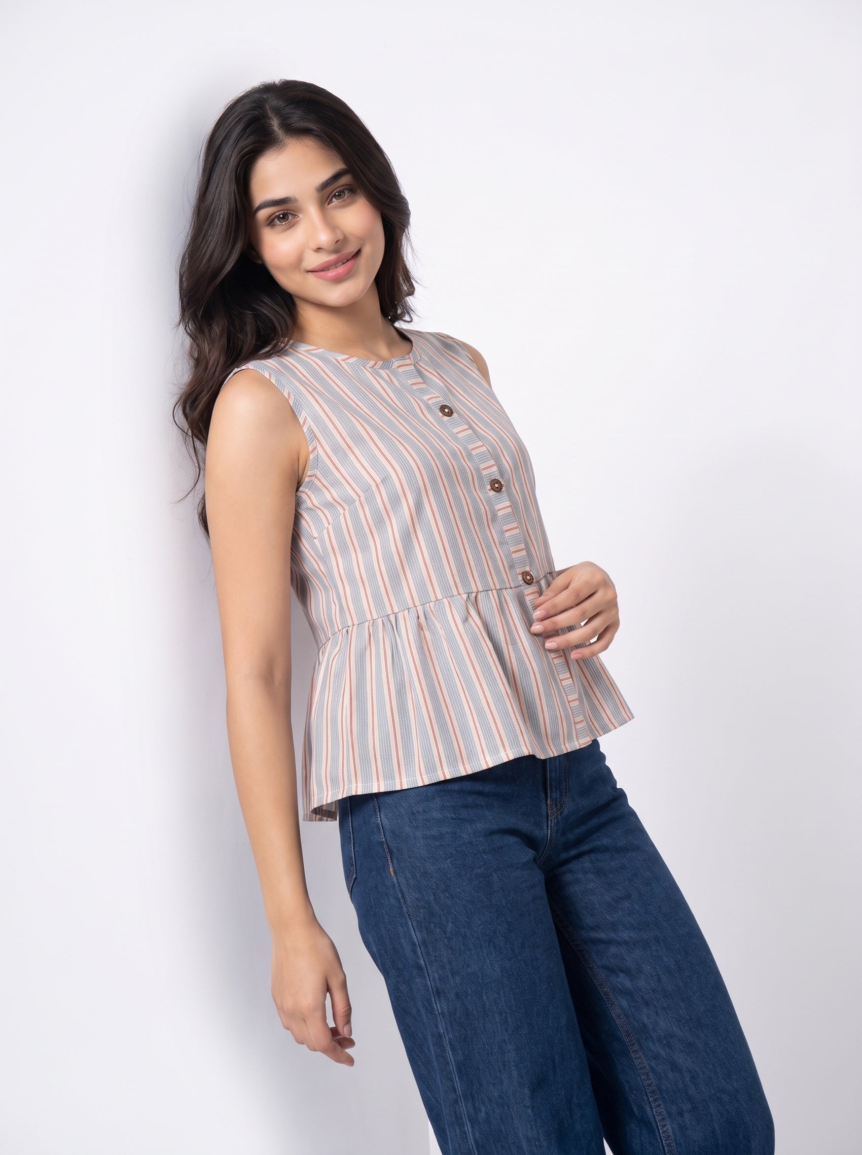 A woman modeling a sleeveless button-front peplum top with vertical peach and blue stripes on a white background, paired with dark blue jeans.