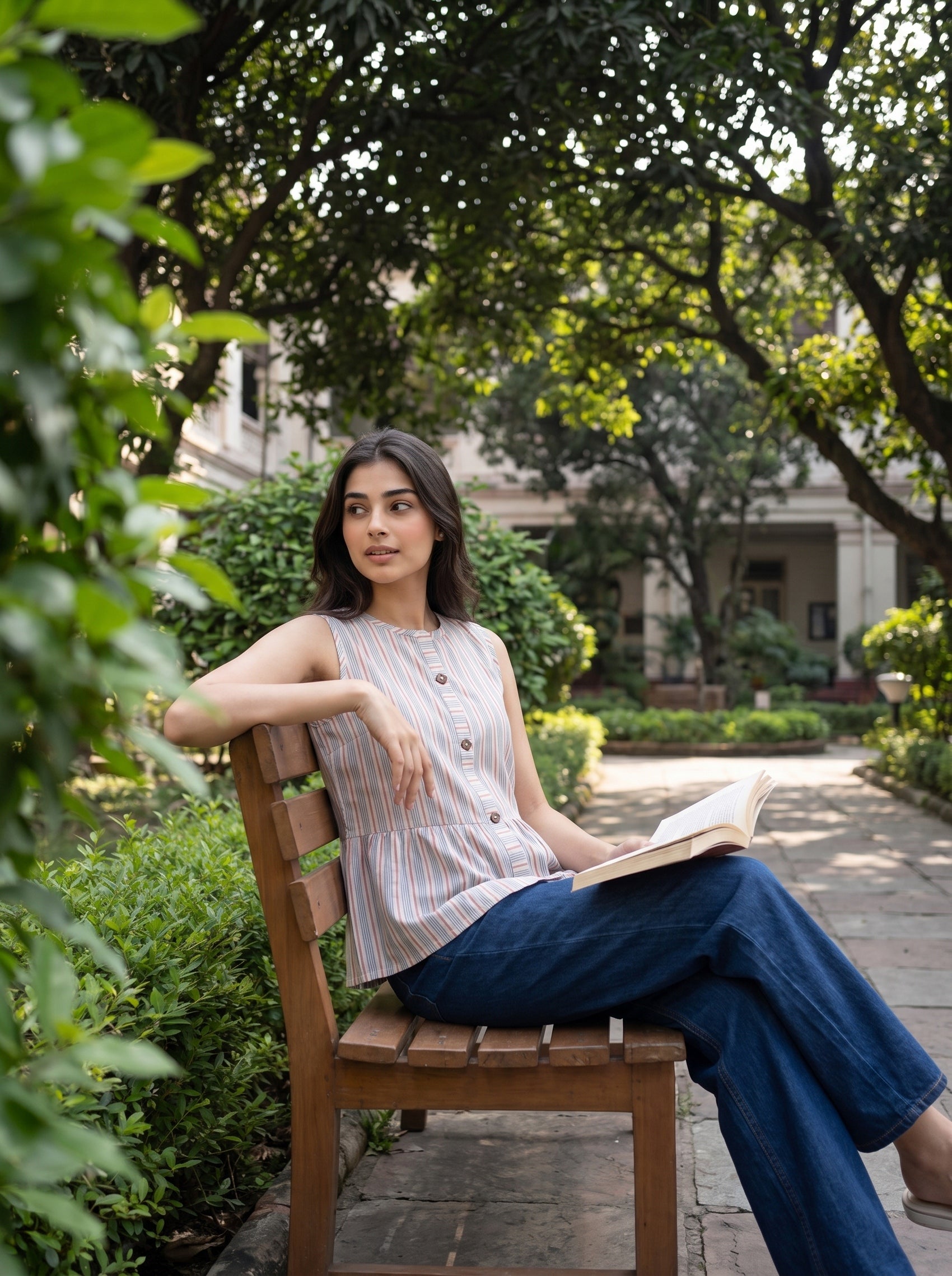 A woman sits on a wooden bench in a garden setting, wearing a sleeveless striped top with buttons and blue wide-leg jeans, holding an open book.