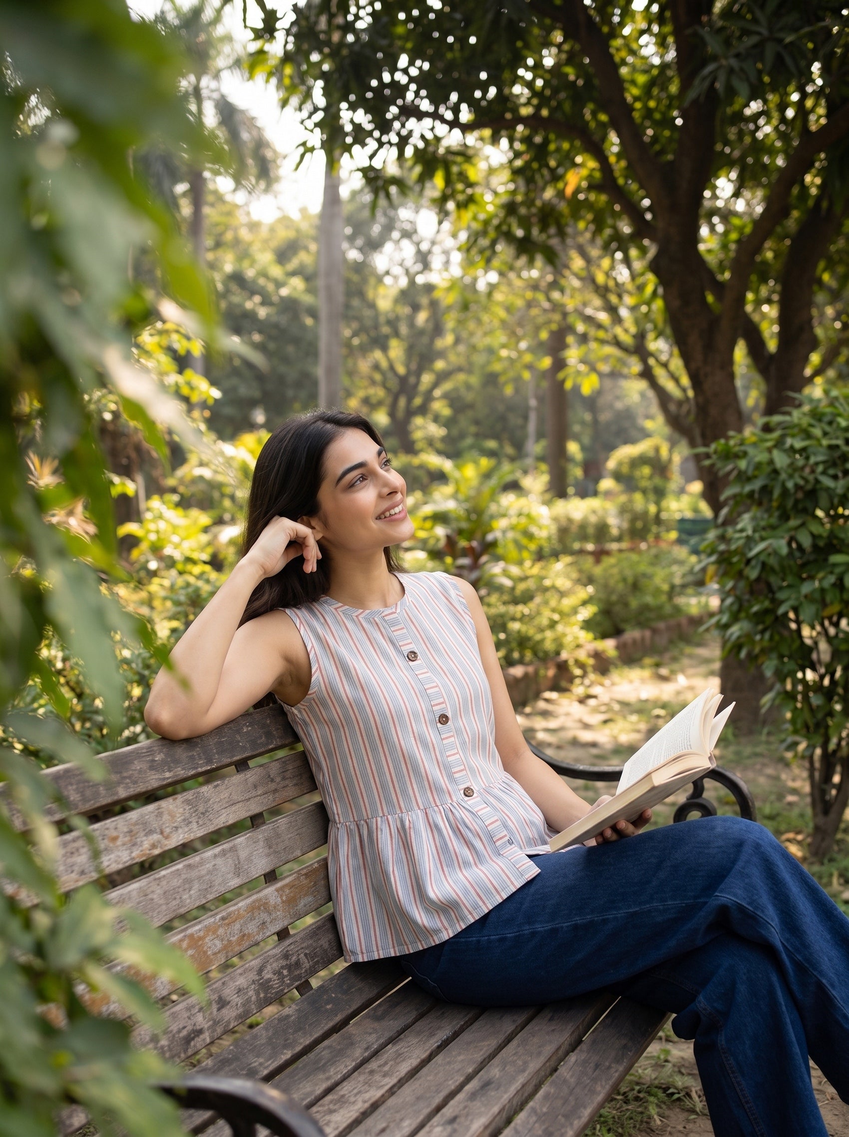 A woman sits on a wooden park bench reading a book, wearing a sleeveless striped top and blue jeans, surrounded by lush green trees and foliage.