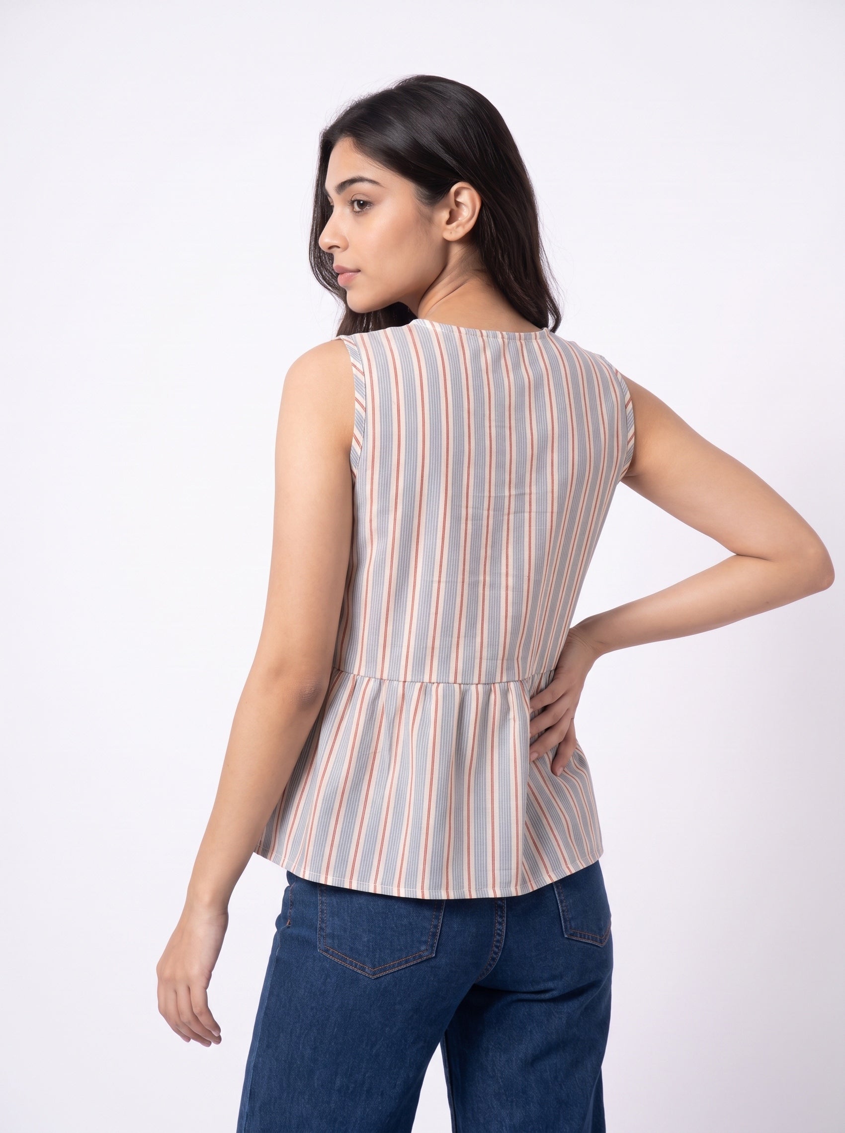 A woman shown from the back wearing a sleeveless peplum top with vertical peach, orange, and cream stripes on a light gray base, paired with blue jeans, against a plain white studio background.