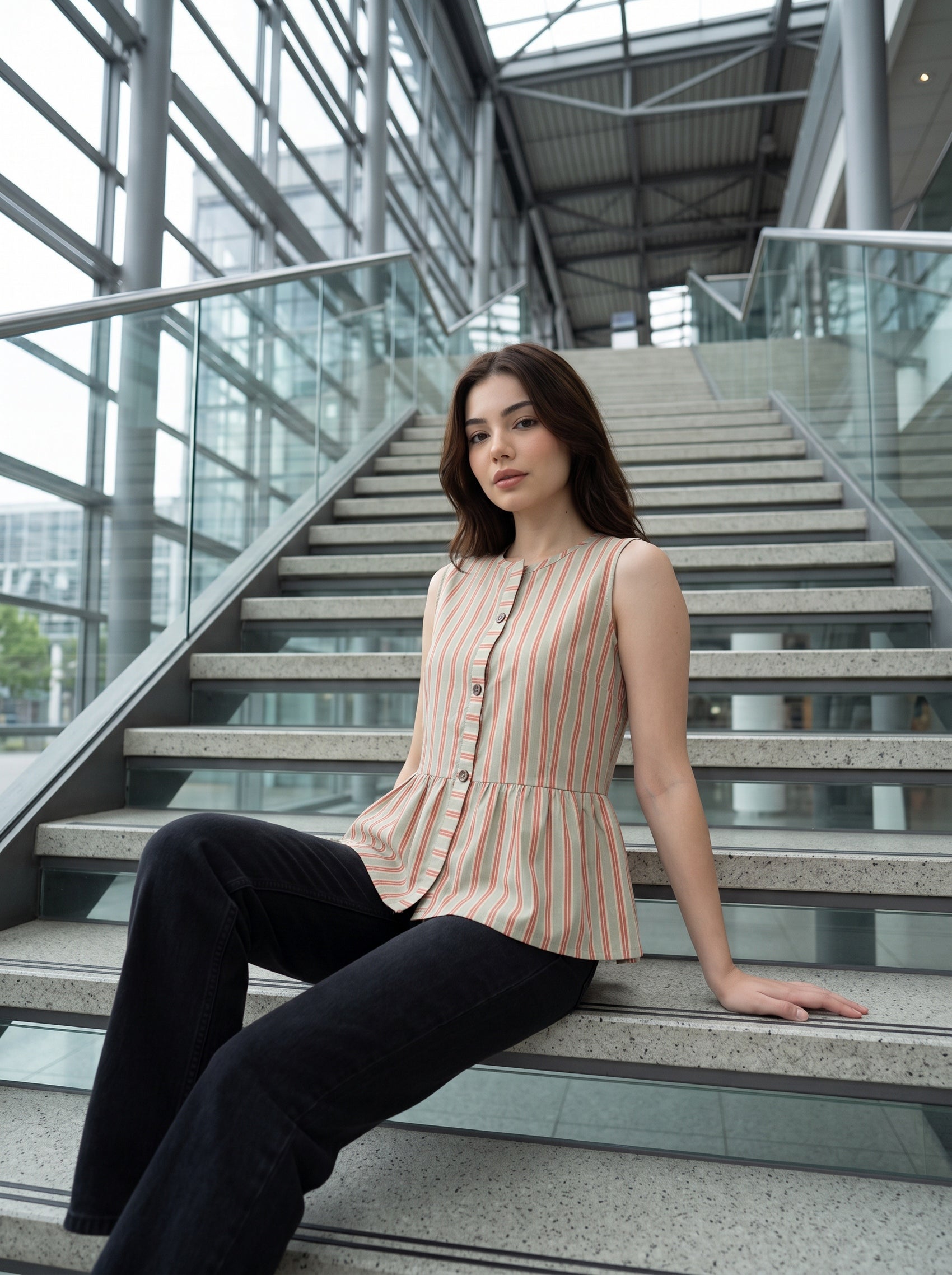 A woman sits on the steps of a modern glass-walled building, wearing a sleeveless, button-front peplum top with vertical peach and dark stripes, paired with black pants.