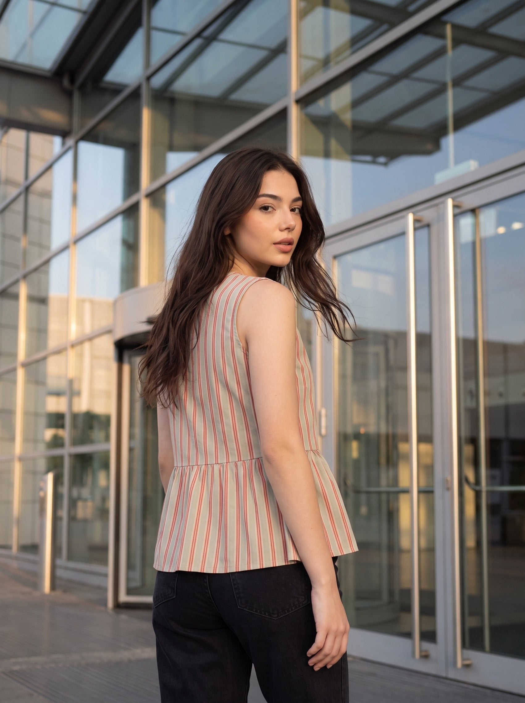 A woman wearing a sleeveless striped peplum top and black pants, standing in front of a modern glass building entrance.