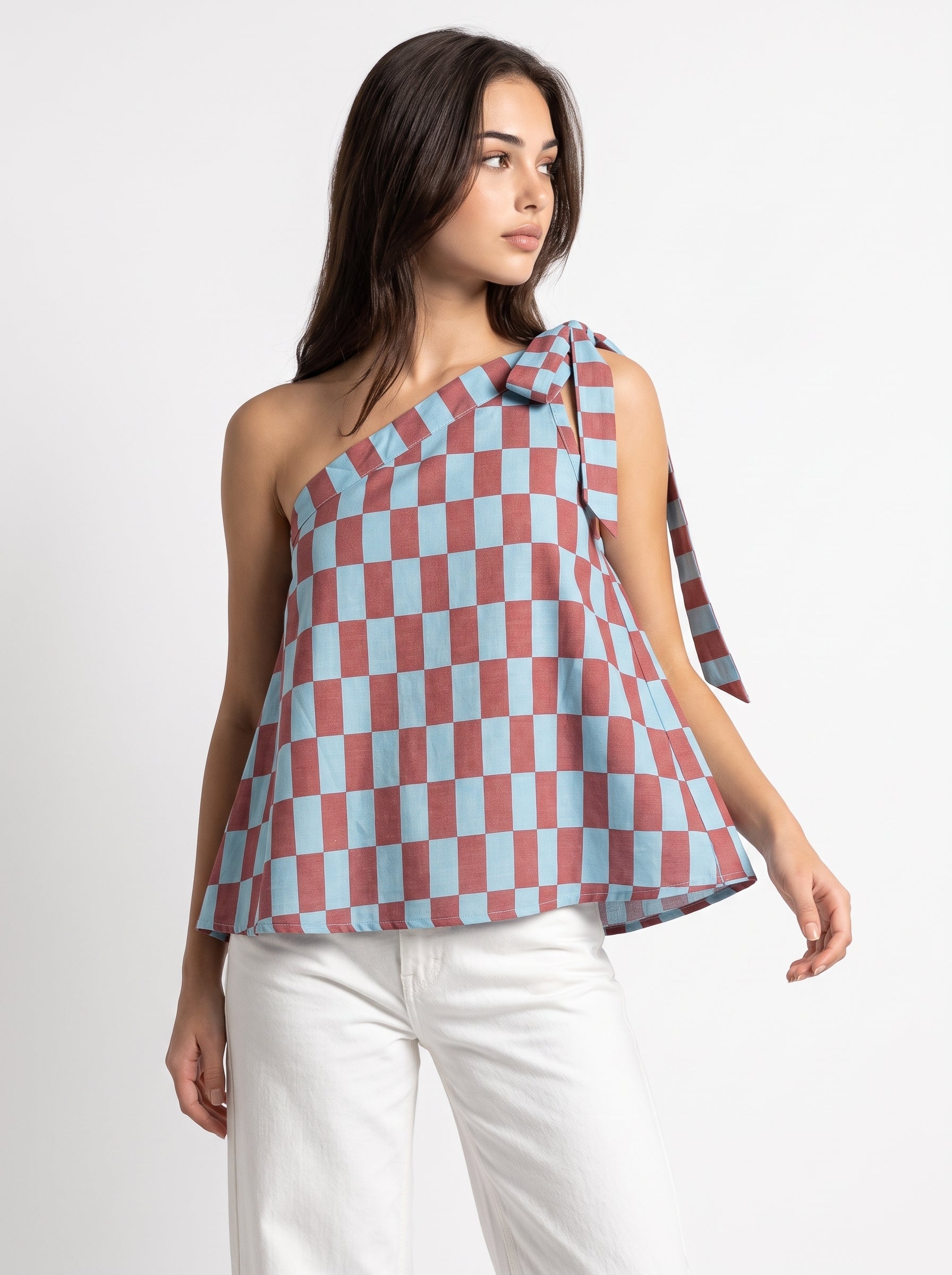 A woman modeling a one-shoulder peplum top with a dusty rose and light blue checkerboard pattern, paired with white pants, against a plain white studio background.