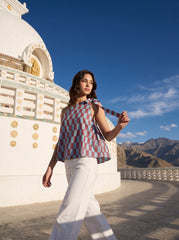 A woman wearing a red, blue, and white checkered sleeveless top and white pants walks in front of a large white stupa with mountains and a blue sky in the background.