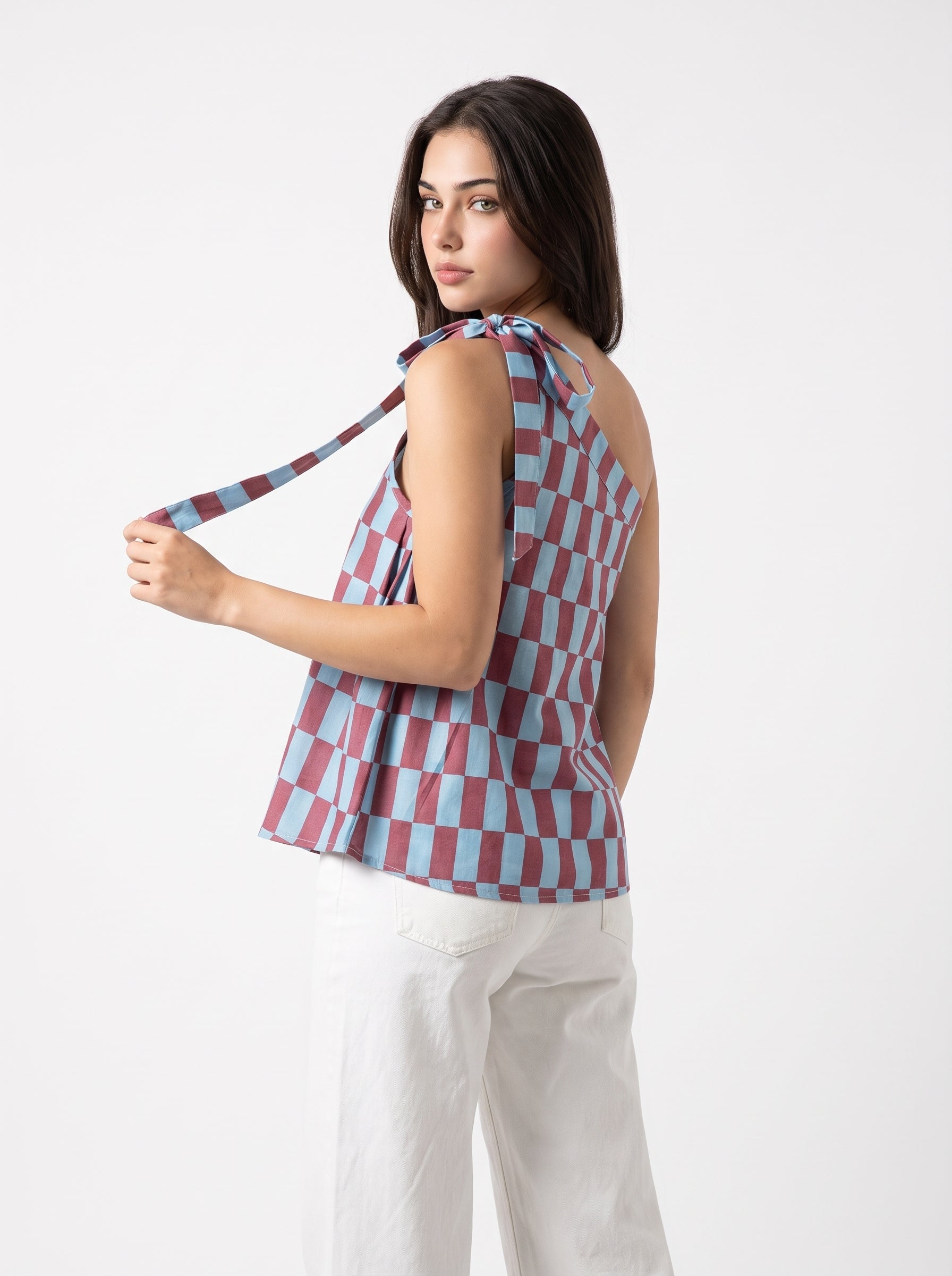 A woman modeling a sleeveless tie-neck top with a pink, blue, and white checkerboard pattern, paired with white pants, against a plain white studio background.