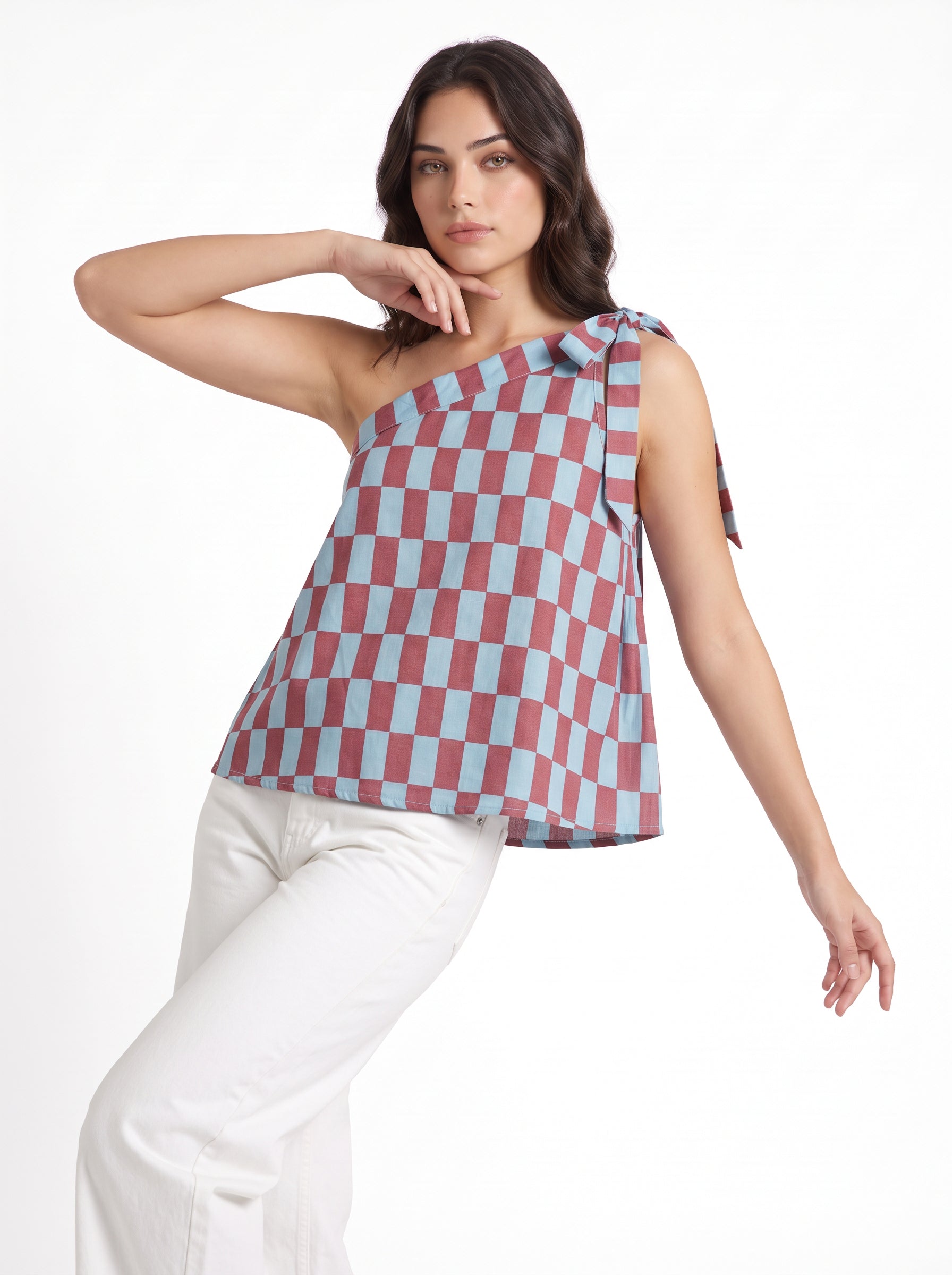 A woman modeling a one-shoulder sleeveless top with a dusty red and light blue checkerboard pattern, paired with white pants, against a plain white background.