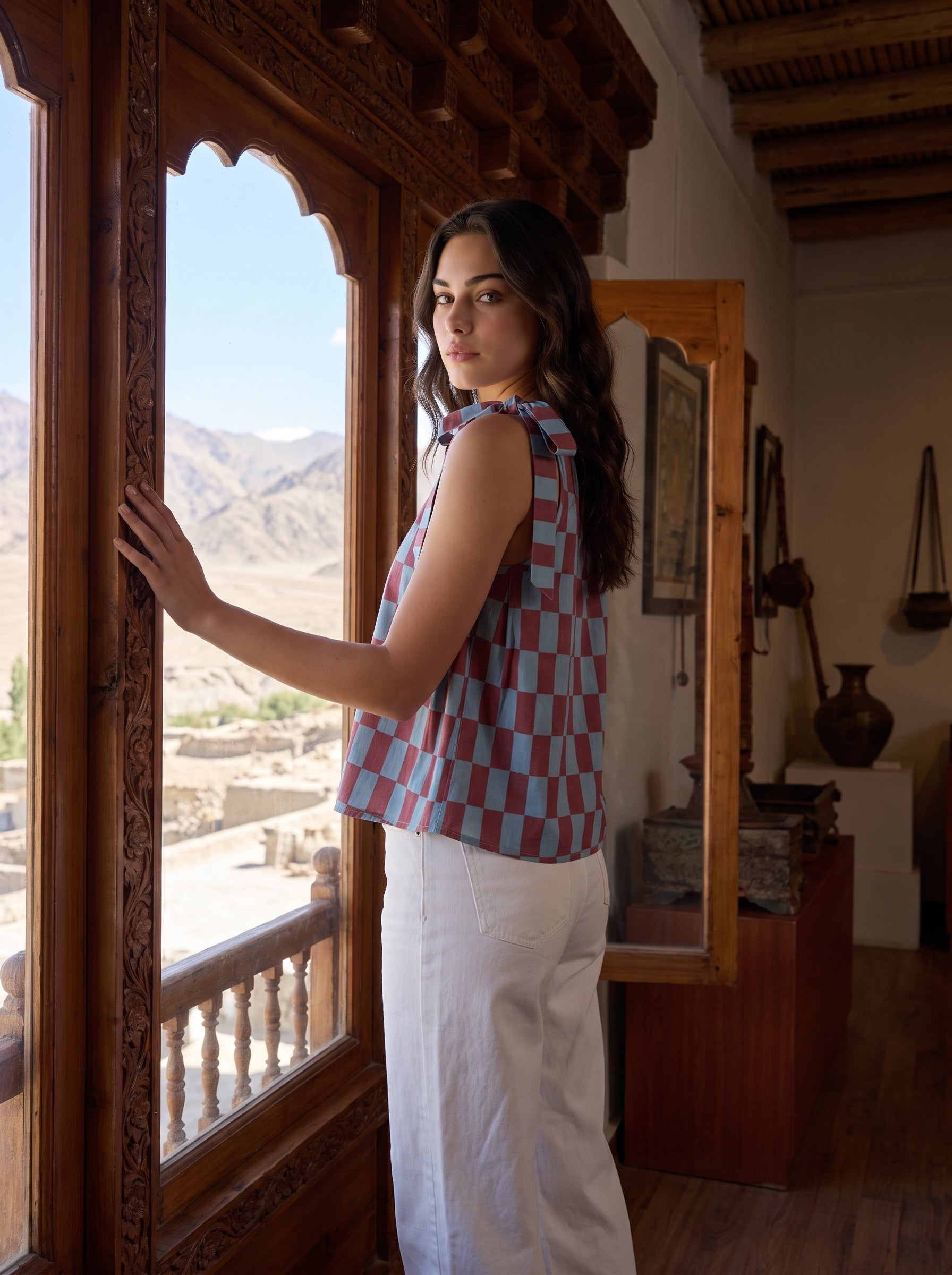 A woman wearing a sleeveless checkered top with blue, pink, and white squares, paired with white pants, standing by a large carved wooden window in a rustic interior with a mountain view.