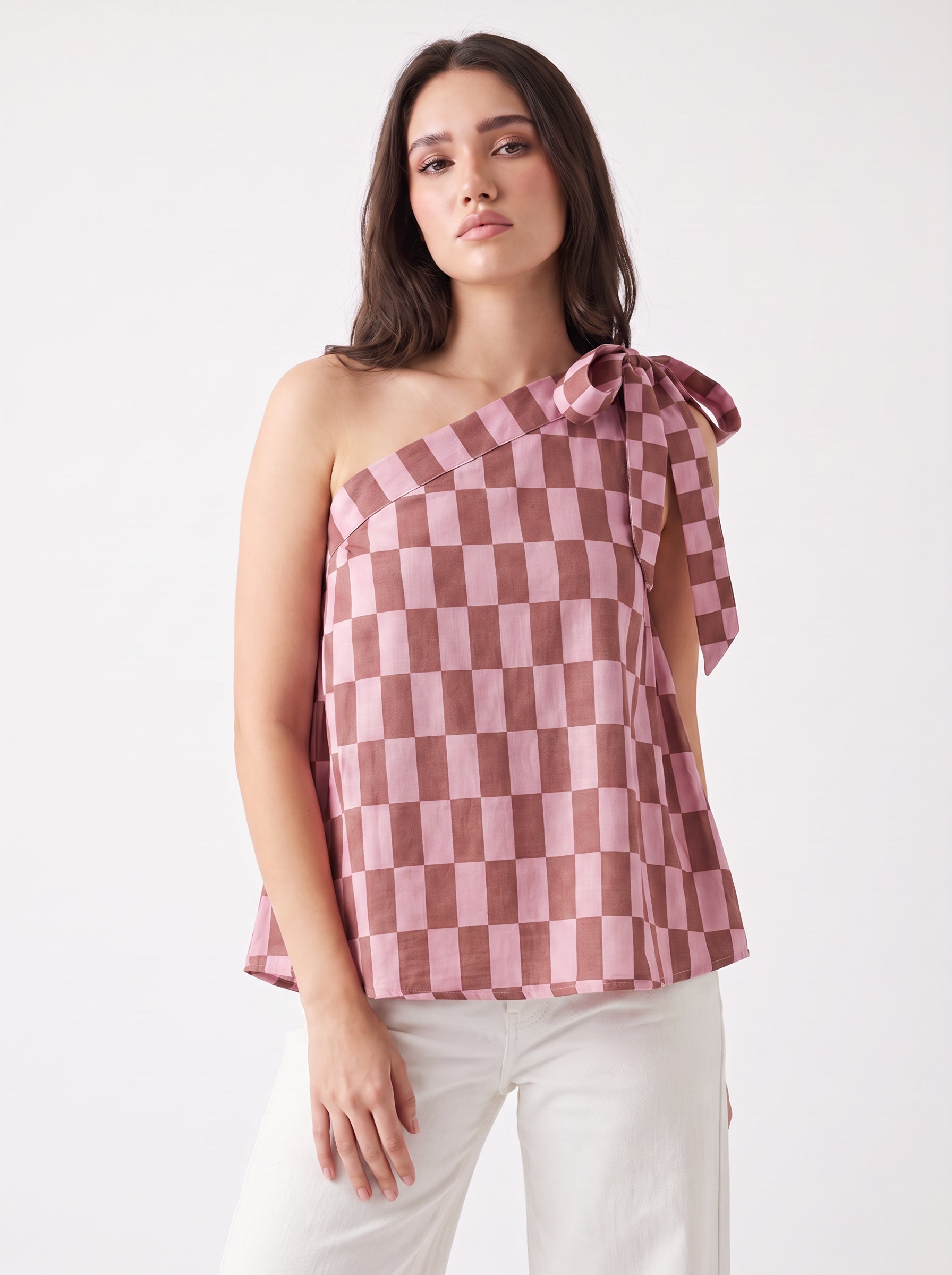 A woman modeling a one-shoulder top with a dusty pink and brown checkerboard pattern and a side bow detail, paired with white pants against a plain white studio background.