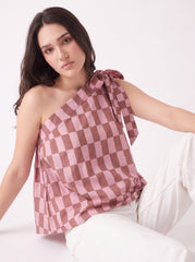 A woman wearing a pink and brown checkerboard one-shoulder top with a side tie, paired with white pants, posed against a clean white studio background.