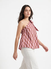 A woman wearing a pink and brown checkerboard one-shoulder top with a side tie, paired with white pants, posed against a clean white studio background.