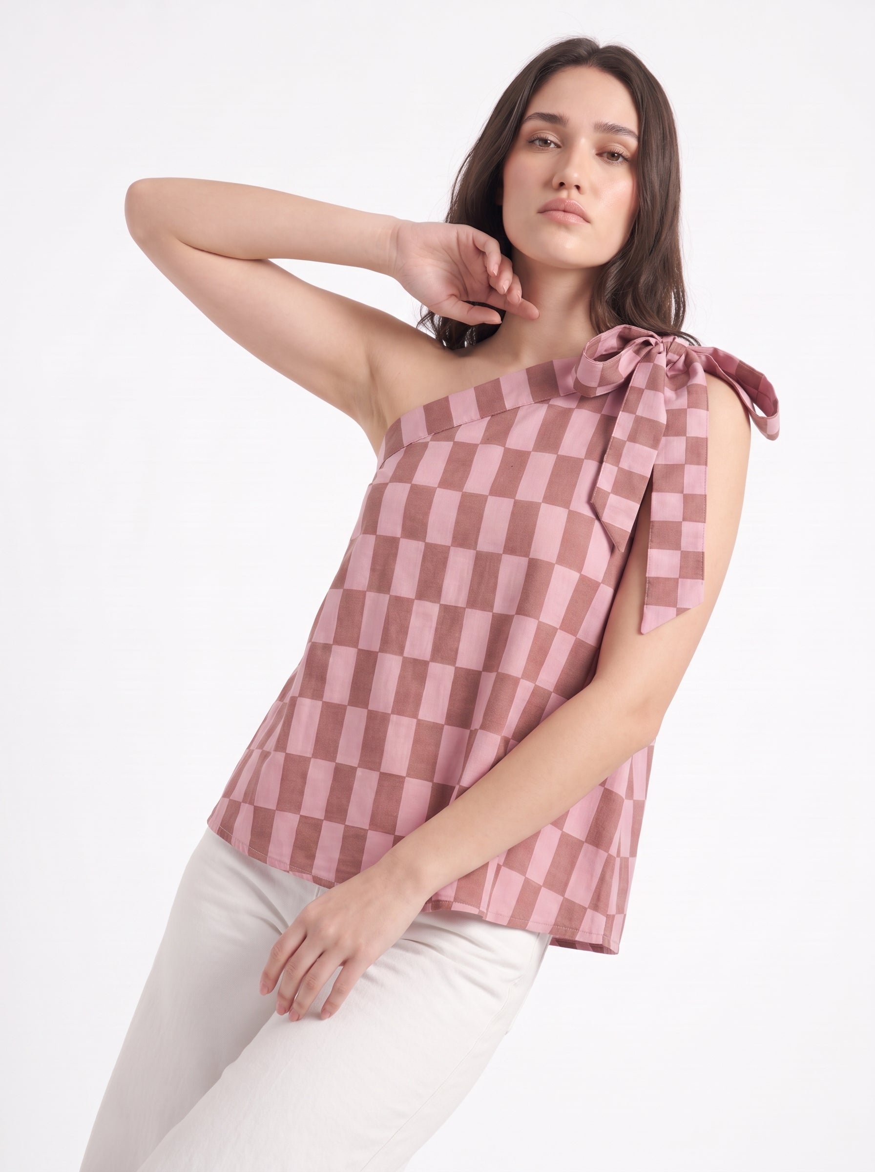 A woman modeling a pink and brown checkerboard one-shoulder top with a side tie detail, paired with white pants, against a plain white studio background.