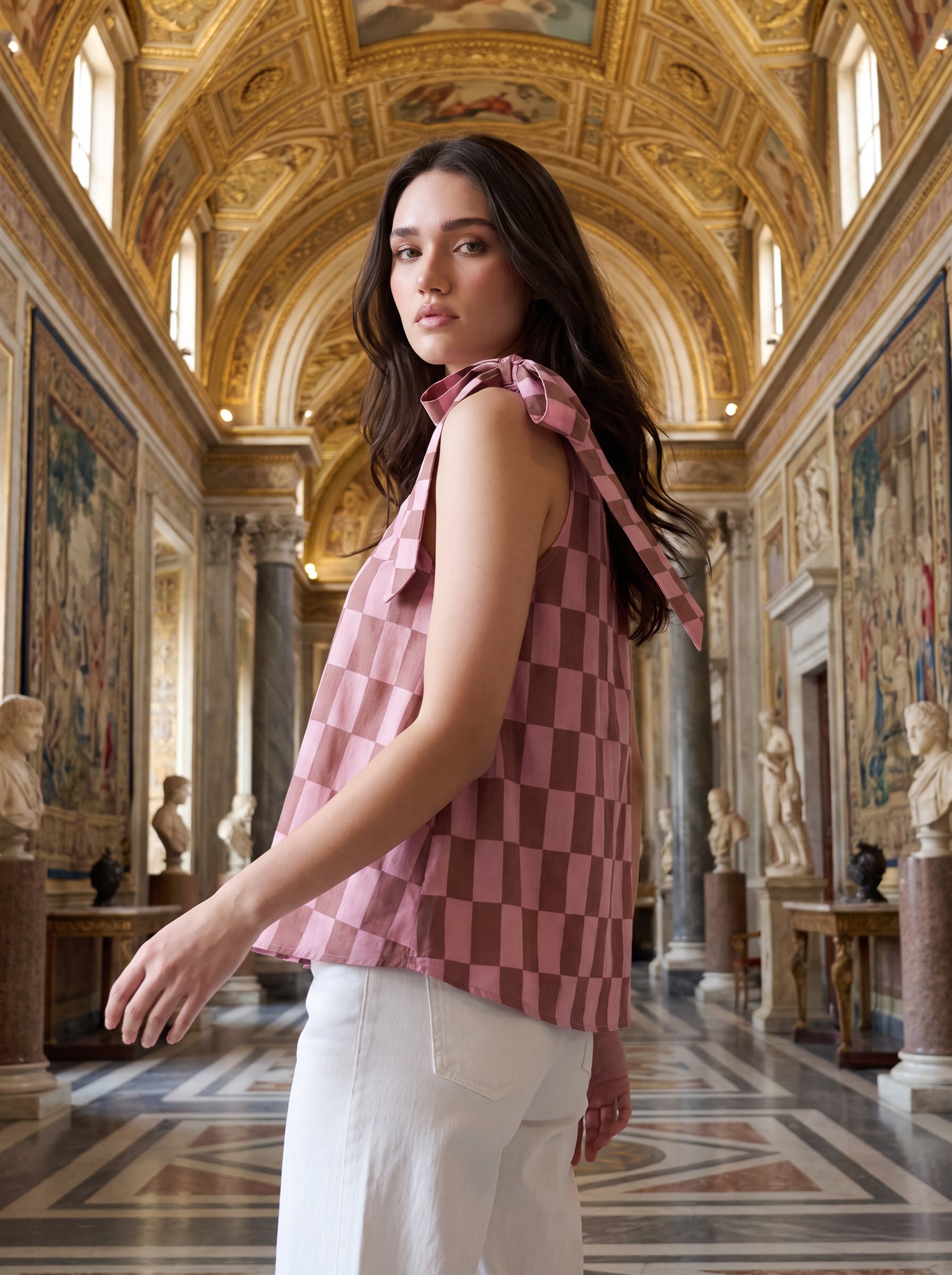 A woman wearing a pink and brown checkerboard one-shoulder top with a side tie, paired with white pants, posed against a clean background.