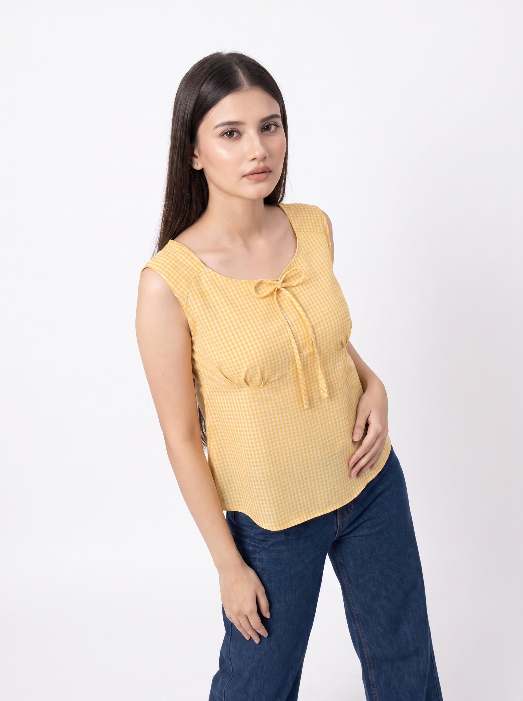 A woman modeling a yellow sleeveless top with a small gingham pattern and a tie detail at the neckline, paired with dark blue jeans, against a plain white studio background.