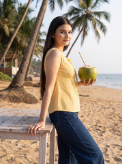 A woman stands on a sandy beach next to a wooden table, holding a green coconut with a straw. She is wearing a yellow gingham sleeveless top and dark blue jeans, with palm trees and the ocean in the background.