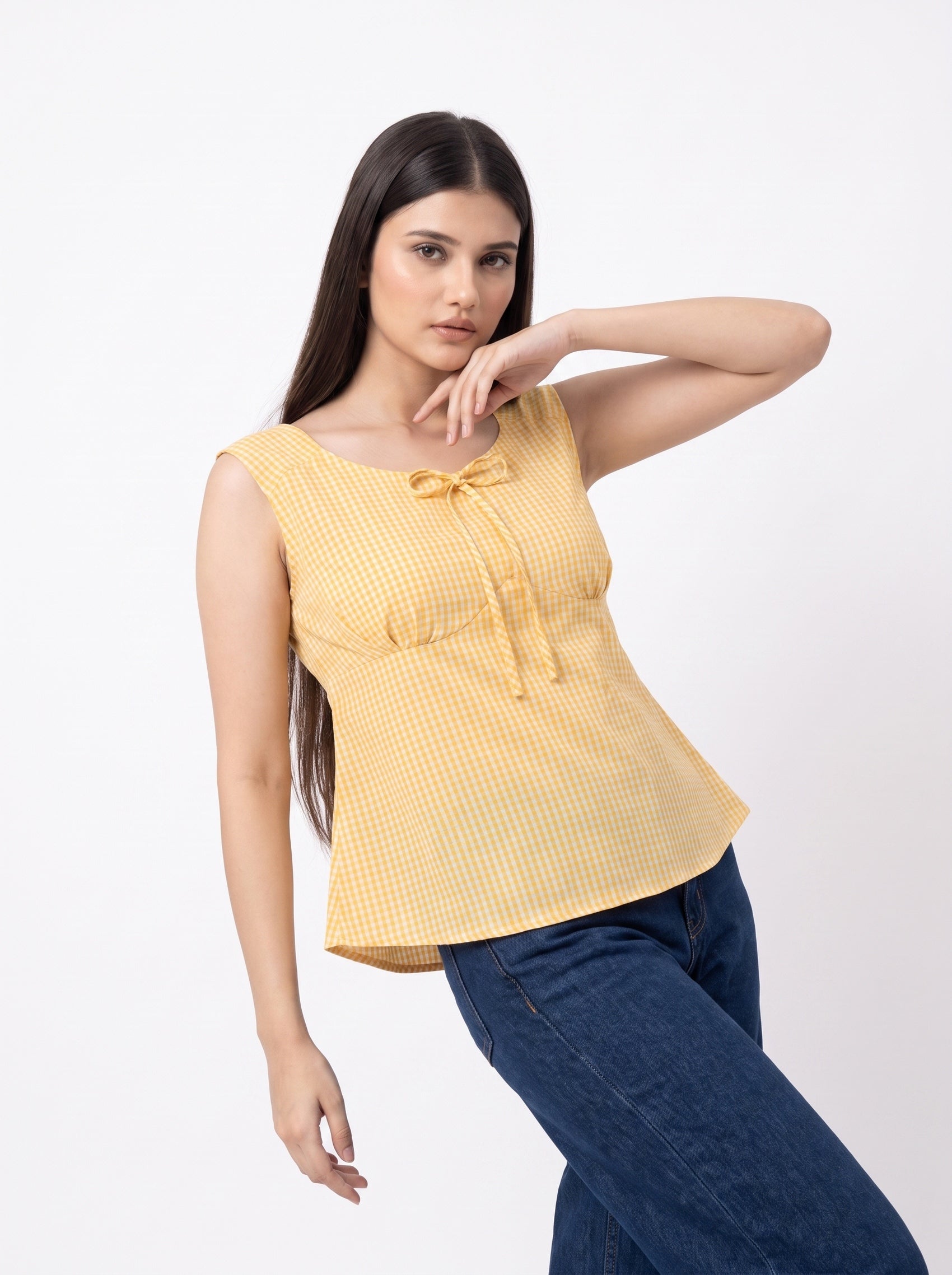 A woman modeling a yellow and white gingham sleeveless top with a tie detail at the neckline, paired with dark blue jeans, against a plain white studio background.