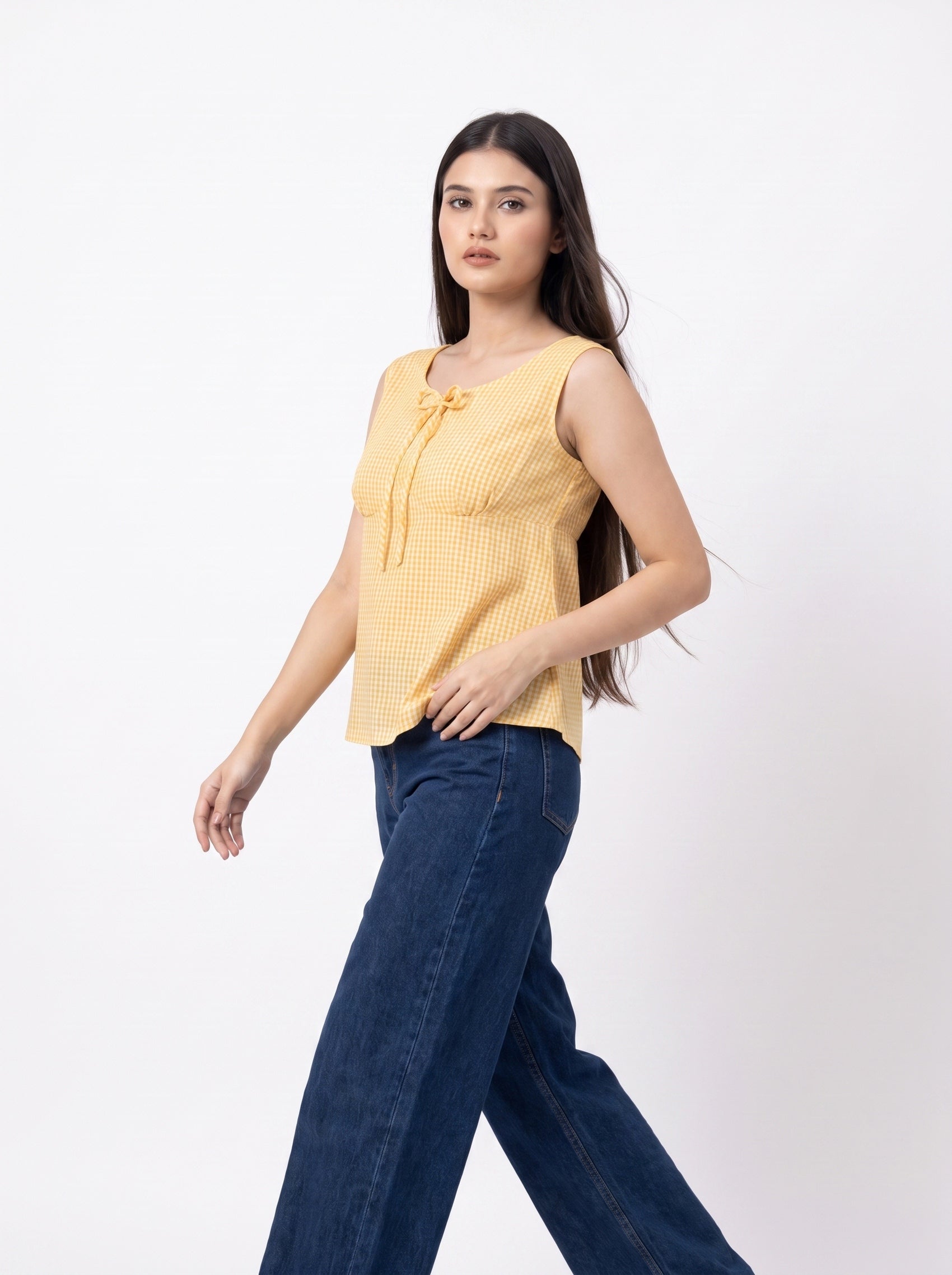 A woman modeling a sleeveless yellow textured top with a tie detail at the neckline, paired with dark blue jeans, against a plain white studio background.
