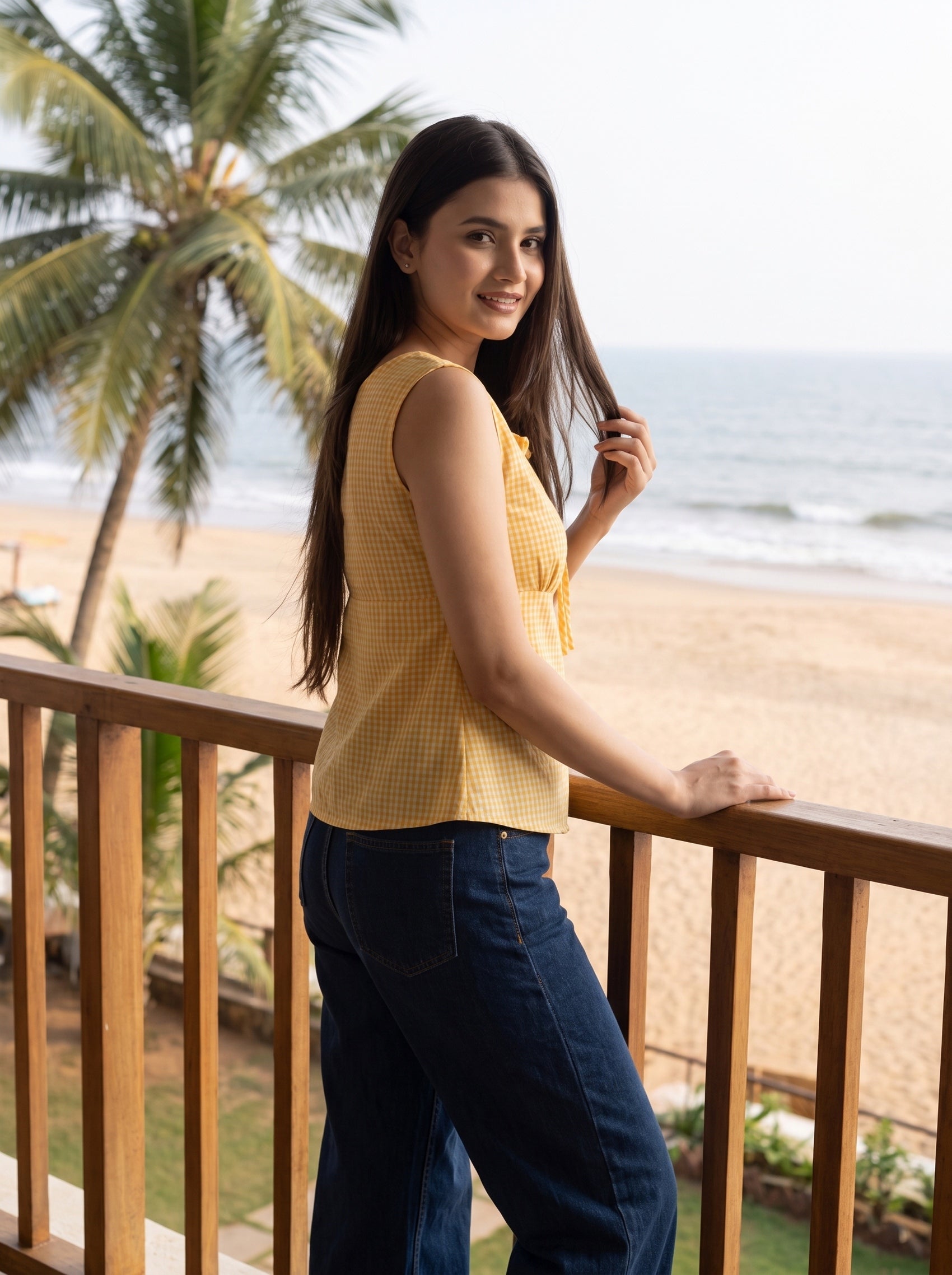 A woman stands on a wooden balcony overlooking a beach with palm trees and the ocean, wearing a yellow sleeveless top and dark blue jeans.