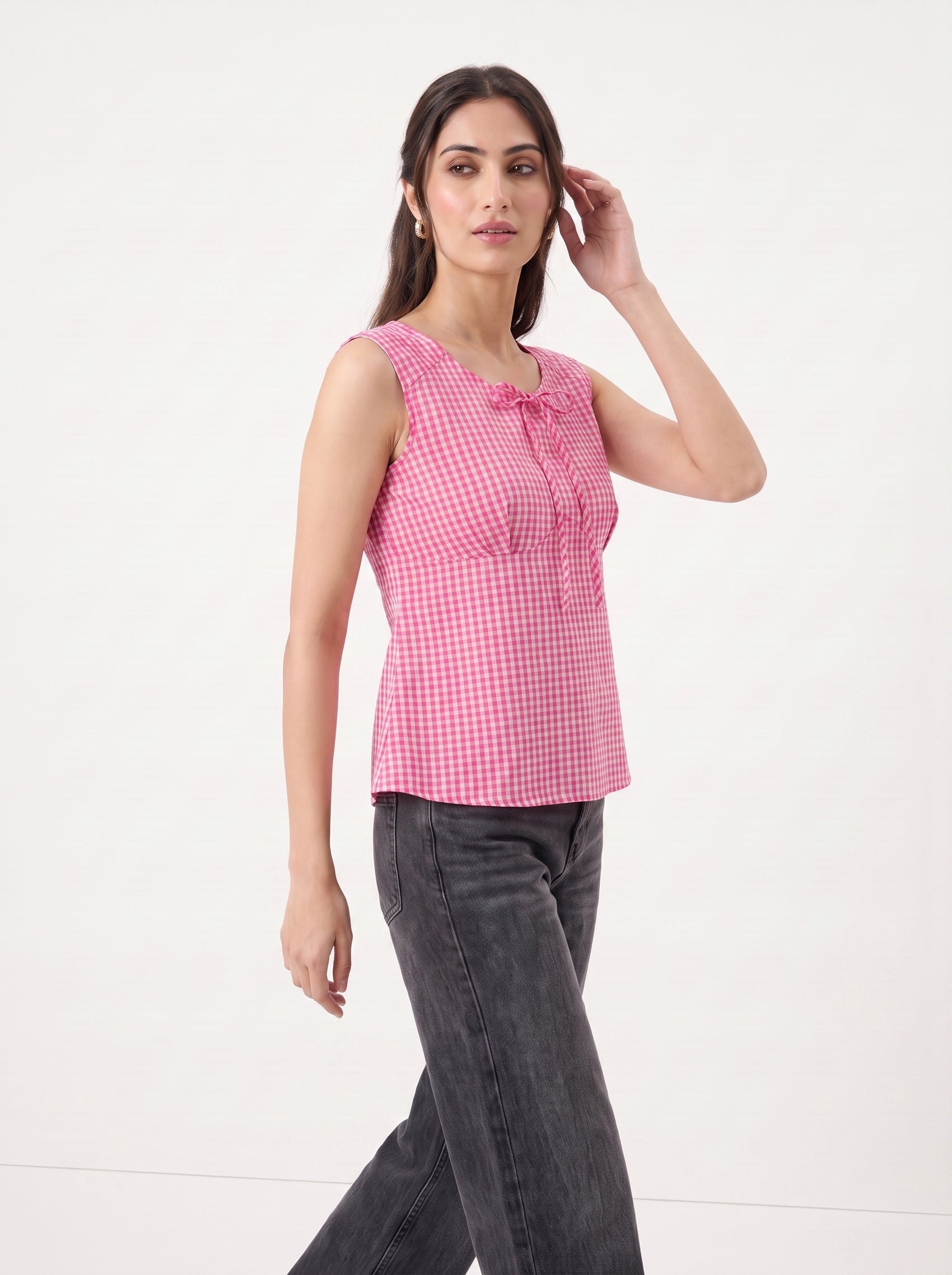 A woman modeling a sleeveless pink and white gingham top with a tie-neck detail, paired with dark gray jeans, against a plain white studio background.